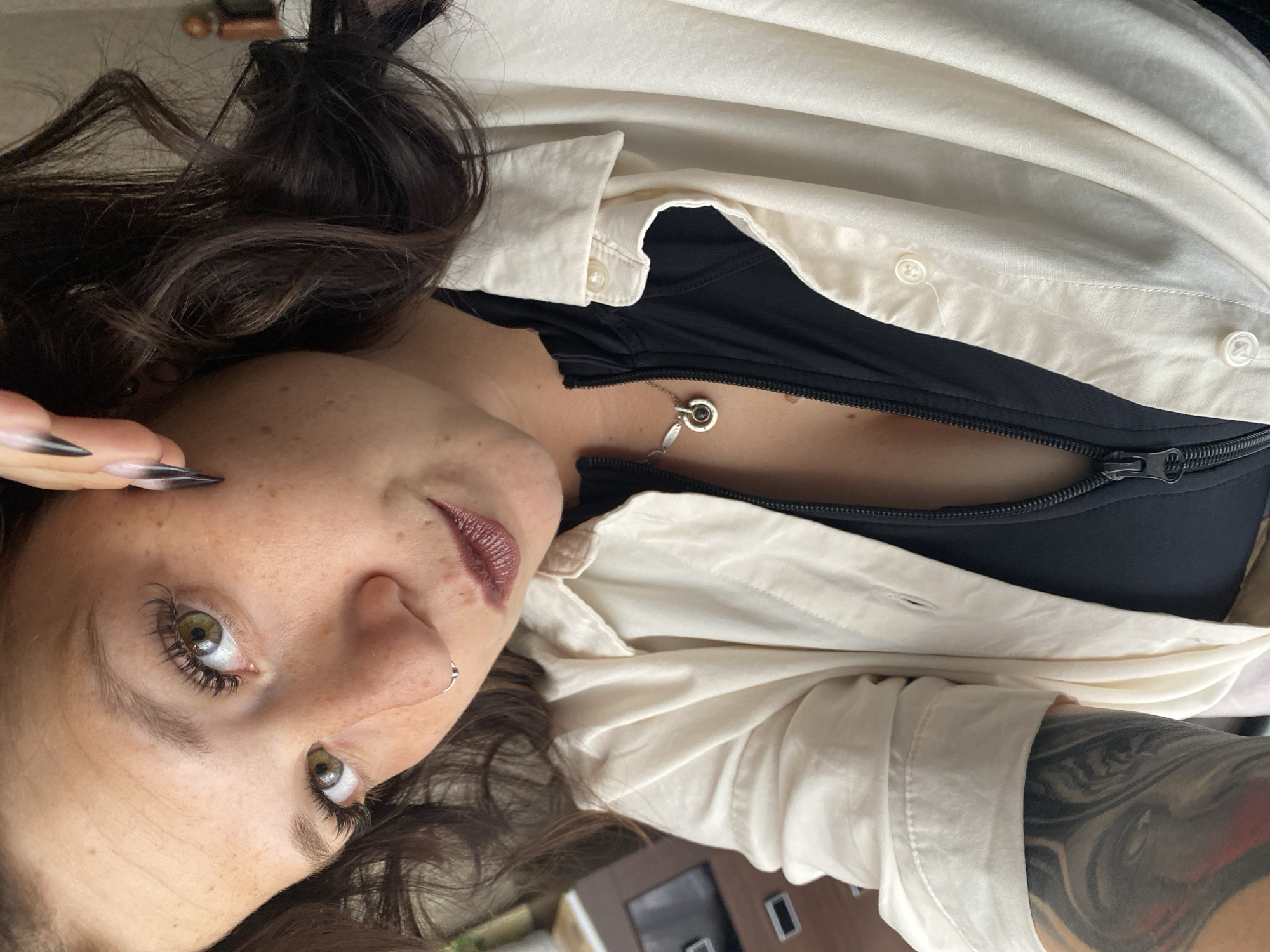 A close-up, selfie-style photograph shows a woman with light brown curly hair, green eyes, and freckles. She is wearing a black zipper-up top and an unbuttoned cream-colored collared shirt over it. A silver necklace with a circular pendant is visible at her throat. Her left arm, which is tattooed, is visible on the left side of the frame. She is wearing dark nail polish on her right hand. The background is blurred and appears to be indoors, possibly an office environment with furniture visible. The image does not contain any elements that would suggest a specific location in Chaikovsky, Russia.