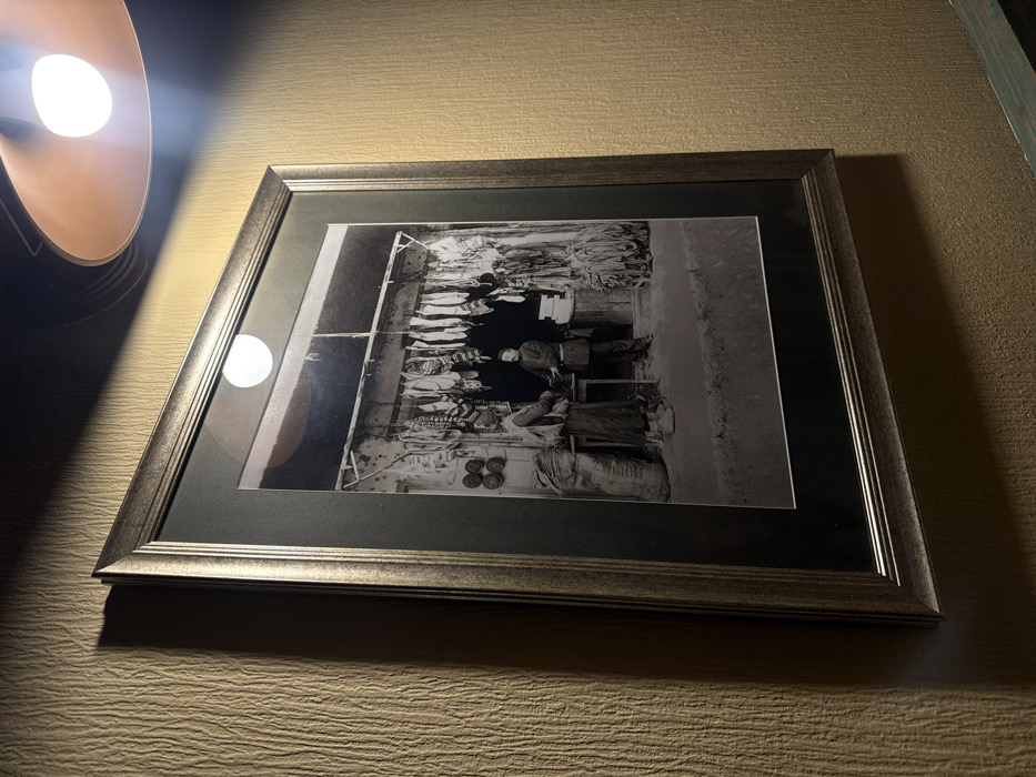 A framed black and white photograph is displayed on a vertically textured, light brown wall. The frame is metallic grey with a slight double-bevel design and a dark matte border. An exposed incandescent bulb in a copper-colored shade hangs in the upper right, casting direct light and reflections on the photograph's glass, notably a distinct circular reflection near the top center of the image.
The photograph depicts a historical street market scene in Tbilisi, Georgia, likely an open-front shop or stall. Two adult males in traditional attire are positioned centrally. On the left, a man, possibly a customer or vendor, wears a long tunic, wide trousers, and a cap, with a large, tall sack leaning against him to his left. He faces right, his hands appearing to engage in an exchange over a small, low table or counter. Opposite him, on the right, stands another man, presumably the shopkeeper, also in traditional dress with a dark cap, interacting with the first individual across the table.
Above the men, numerous items, consistent with cured meats or sausages, hang from a horizontal bar extending across the width of the stall. To the left of these hanging goods, four circular objects are affixed to the wall. To the right, various ropes, braids, or bundles of dried goods are hung and stacked. The ground in the foreground of the photograph is unpaved, appearing as dirt or a rough path. The background within the stall is dark, indicating a shadowed interior or opening. The overall atmosphere conveyed is that of a bustling, traditional marketplace from an earlier era.