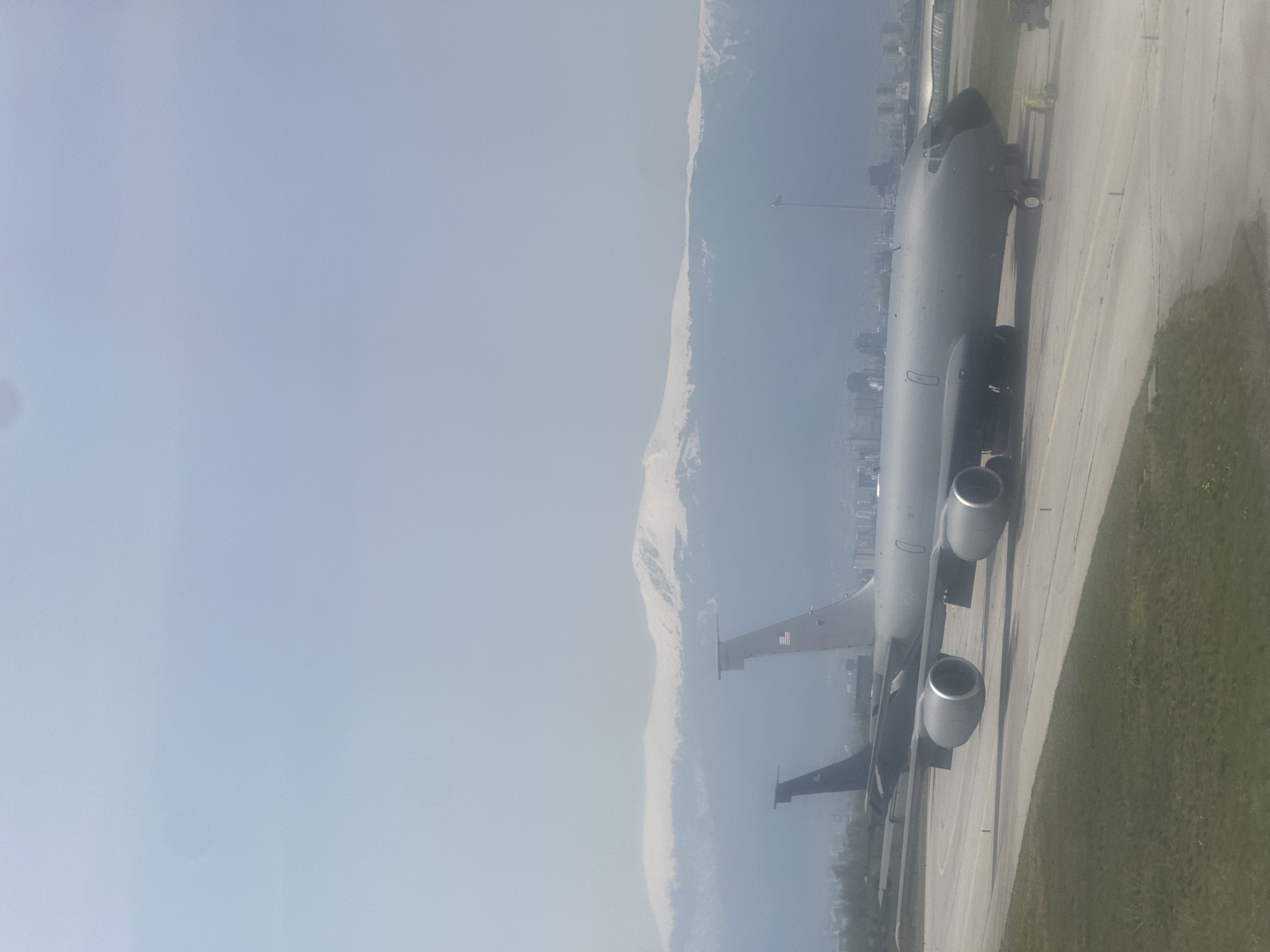 The image shows a KC-135 Stratotanker aircraft on a runway, likely at Sofia Airport, with the Vitosha mountain range visible in the background. The aircraft is positioned on the right side of the frame, with the runway and some grassy areas alongside it. The left side of the image is dominated by a hazy sky and the snow-capped Vitosha mountain. In the distant background, a city skyline is visible, indicating the city of Sofia, Bulgaria. The scene appears to be taken during the day, with overcast or hazy conditions affecting visibility.