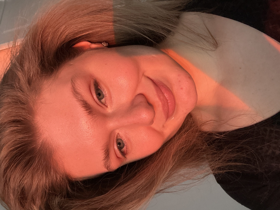 A close-up portrait features a light-skinned female individual in an indoor setting, identified as Kazan, Russia. The subject has light blonde hair, blue-green eyes, and a slight, closed-mouth smile. Her face is predominantly illuminated by a strong, reddish-orange light source from the viewer's right, creating a distinct diagonal shadow line across her lower left face and neck. Her right cheek, forehead, and hair are bathed in this warm light. A small, circular stud earring is visible in her right ear. Minor skin irregularities are present on her chin and right cheek. She is wearing a dark, V-neck garment, visible at the bottom of the frame. The background is an indistinct, light-colored surface.