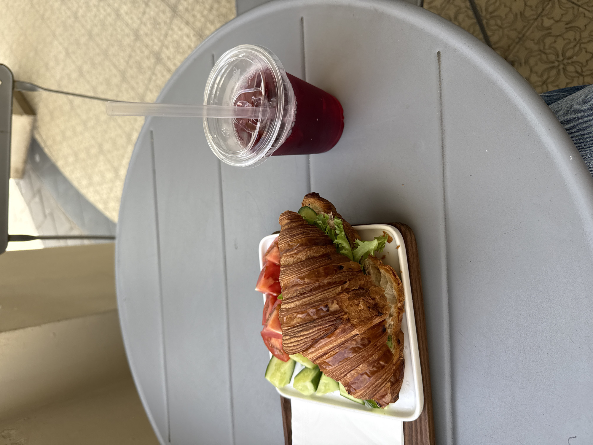 The image captures an outdoor dining scene, likely in Tel Aviv, Israel, as suggested by the city's urban characteristics. A grey, round table occupies most of the frame, upon which a croissant sandwich and a red drink are placed.
The croissant sandwich is served on a small, rectangular white plate with a wooden frame. It contains sliced tomatoes, cucumbers, and lettuce visible around the croissant.
The red drink, possibly juice or a mixed beverage, is in a clear plastic cup with a domed lid and a straw extending out.
The table is situated outdoors, indicated by the visible tiling on the floor and a glimpse of seating beside the table. A portion of a person wearing denim is visible on the edge of the table, suggesting the setting is a casual outdoor café or restaurant. The image is shot from a high-angle perspective.