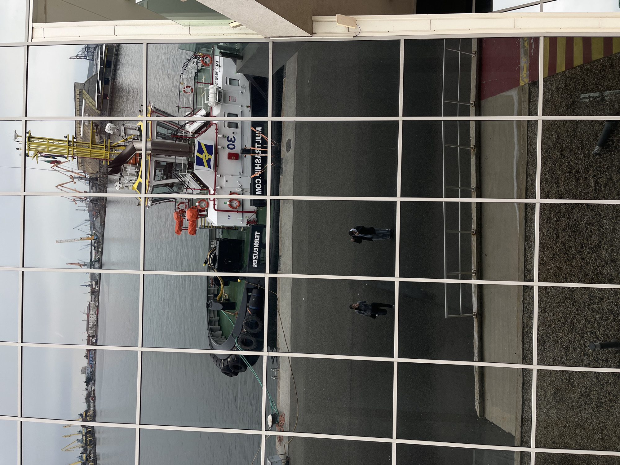 The image is a reflection in a window depicting a scene at the Port of Burgas, Bulgaria. In the reflection, a harbor tugboat named "Teknesensen" with the number 30 is docked. The tugboat has the Swedish flag on its side. In the background of the tugboat, across the water, is the skyline of Burgas, including buildings and cranes. On the right side of the reflection is the quay where the tugboat is docked, with two people walking along it. The window reflection is divided into a grid by the window frames.