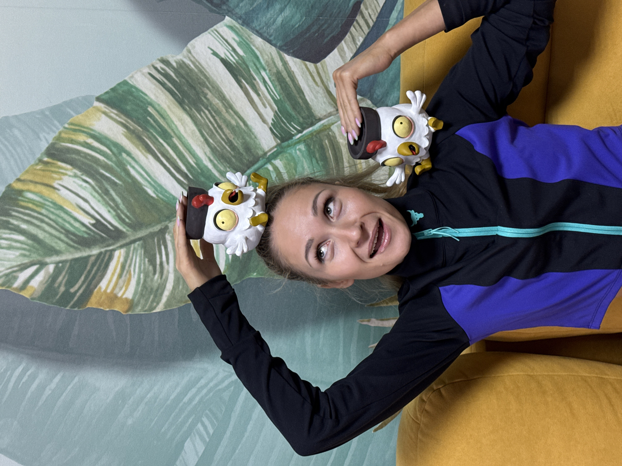 A young woman with blonde hair is posing for a photo on a yellow couch. She is wearing a black and purple athletic jacket with a turquoise zipper. Two cartoonish figurines of white chickens wearing black hats and red combs are placed on her head and shoulder. The background is a wall with a tropical-themed wallpaper featuring large green and yellow leaves. The woman is looking up and to the left with a surprised expression. The scene is likely an indoor setting, possibly a living room or studio. There is no identifiable information about the city of Chaikovsky, Russia.