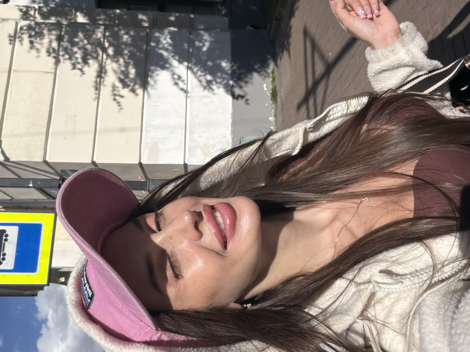A young woman with long brown hair, wearing a pink baseball cap and a cream-colored fleece jacket, poses for a selfie outdoors. Her eyes are closed, and she has a slight smile. She is wearing a dark brown top and a delicate necklace with a small pendant. A bus stop sign with a blue and yellow design is visible in the background, suggesting an urban setting. The architecture in the background appears to be a building with light-colored siding and horizontal banding. Shadows of trees are cast across the building and the ground, indicating a sunny day. The ground is paved with brick-like patterns. The overall scene is likely a street or sidewalk in a city.