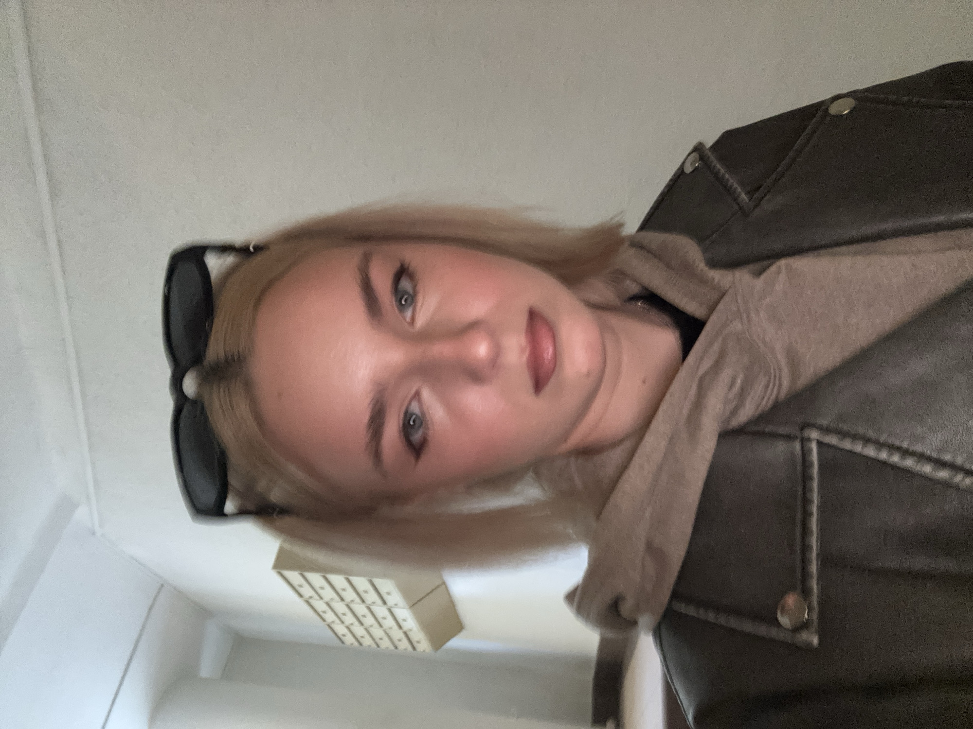 A young woman with blonde hair styled in pigtails and sunglasses on top of her head is shown in a medium shot. She is wearing a dark leather jacket over a beige hoodie. The background is a lightly textured wall with a cream-colored mail sorter mounted on it. The lighting is bright, casting a pinkish hue on her face. The location appears to be indoors, possibly a hallway or stairwell.