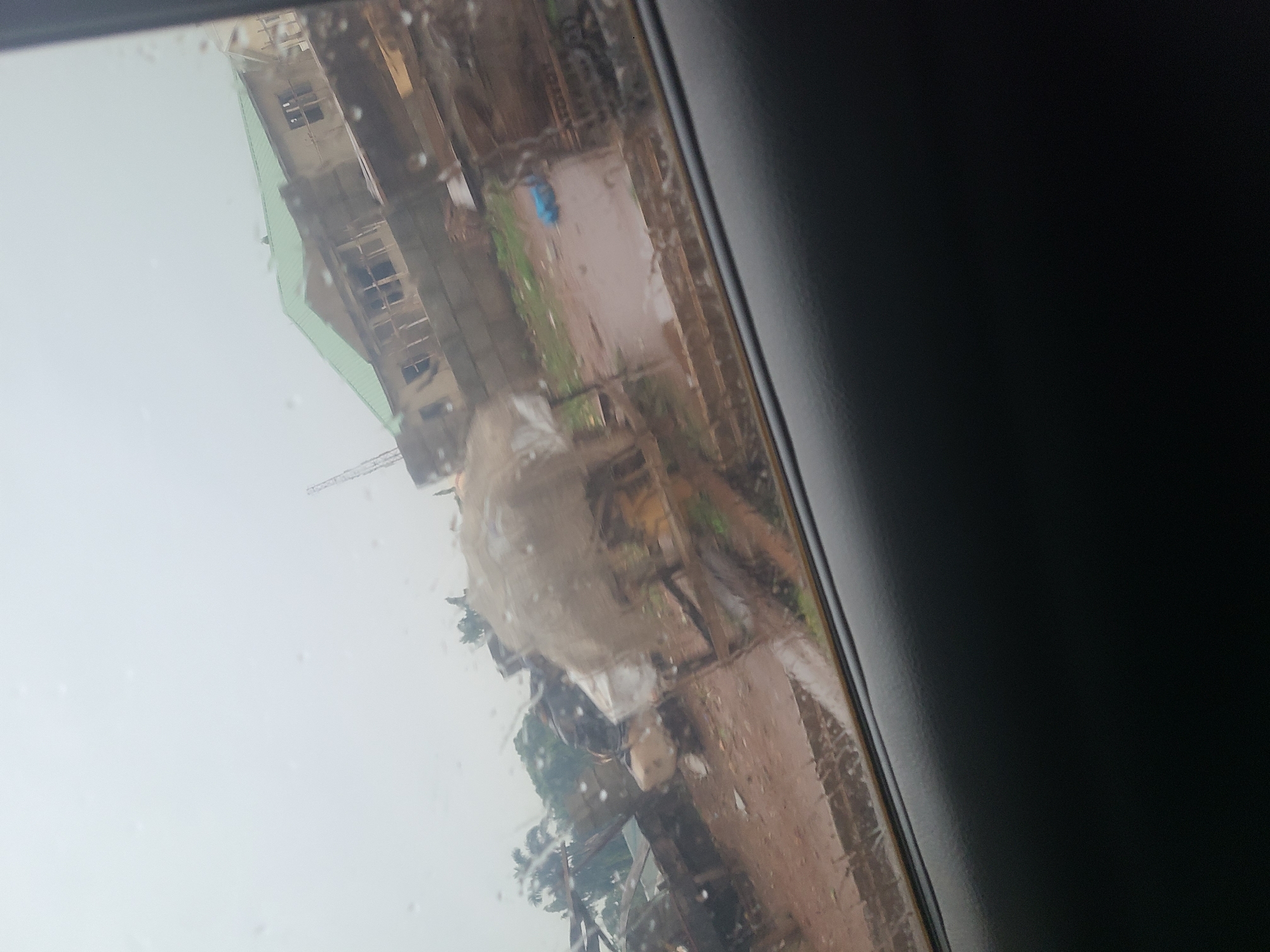 The image shows a view through a car window, with water droplets obscuring the scene outside. In the background, a building with a green roof is under construction, featuring unfinished walls and window frames. To the left of the construction site, there are more structures, some appearing to be temporary shelters or shacks. The ground is muddy and uneven, suggesting an unpaved area. Various objects, including sacks or tarpaulins, are visible, along with some scattered debris. There are no people clearly visible in the image. The overall impression is of a developing or somewhat disorganized urban environment, likely in Karu, Nigeria, given the context. The sky is overcast and hazy.