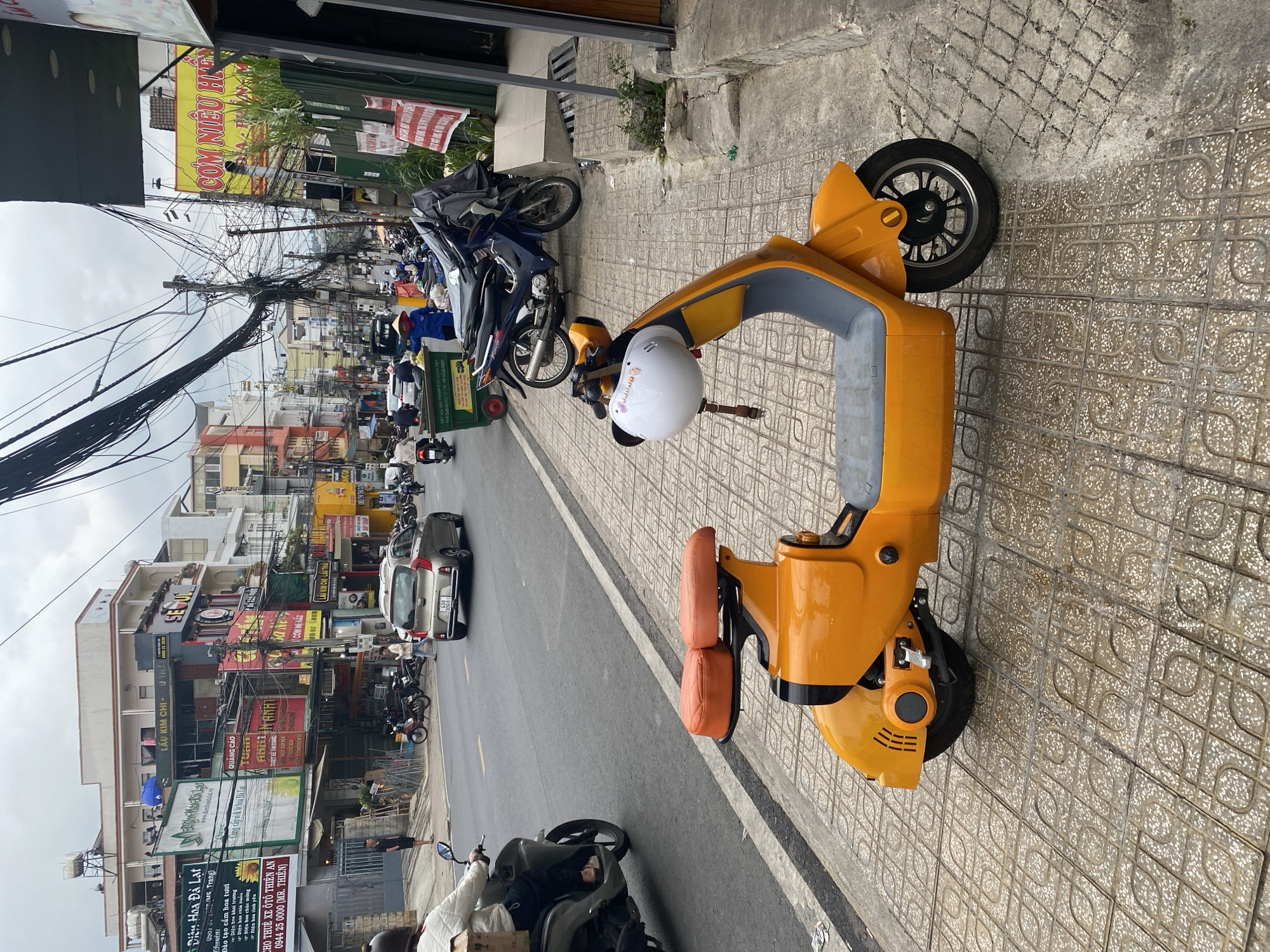 The scene depicts a street in Da Lat, Vietnam. A bright orange electric scooter is parked on the tiled sidewalk. A white helmet rests on its seat. The street is lined with buildings featuring numerous signs in Vietnamese. Above the buildings, a dense network of overhead wires is visible. A car and several motorbikes are moving along the road. There is evidence of retail businesses with signs indicating services for flowers and automotive rentals. The architecture reflects a mix of traditional and modern Vietnamese styles. The sky is overcast.