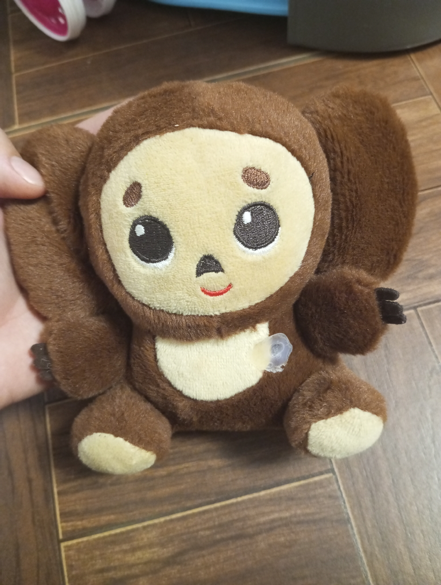 A close-up, waist-up, eye-level shot shows a brown Cheburashka plush toy sitting on a wooden floor. The toy is positioned centrally with its head slightly tilted. Its large, round ears are spread out to the sides. The toy has a light brown, oval-shaped face with large black eyes, two small brown dots above the eyes, and a small red mouth. A light brown oval shape is present on its chest. A small, clear plastic tag is attached to the toy's chest. A human hand is visible on the left side of the image, gently holding the toy's left ear. The background consists of a wooden floor with a visible wood grain pattern. The toy is located in an indoor setting, in the city of Verkhnyaya Pyshma, Russia.