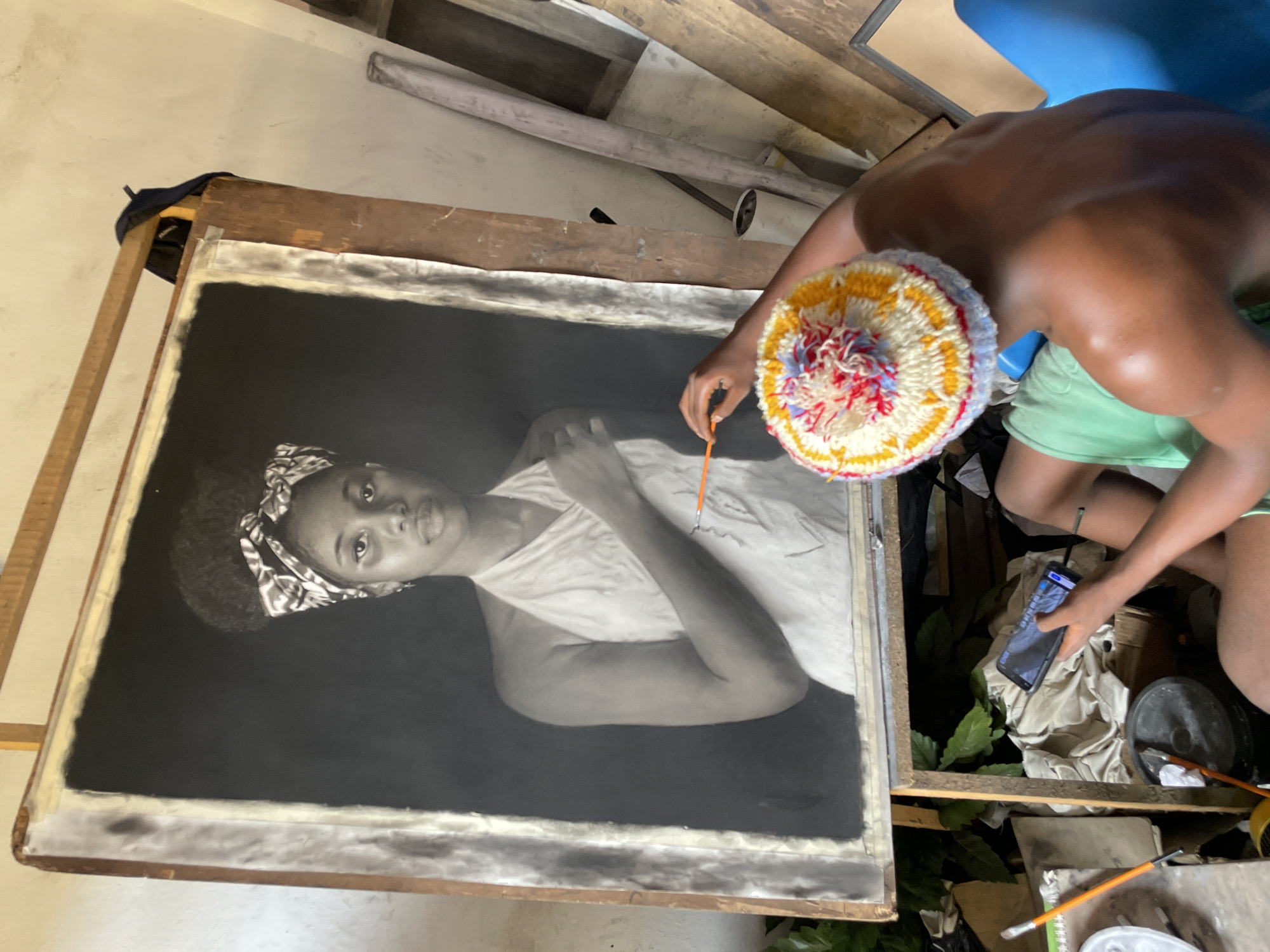 The image shows an artist working on a charcoal portrait of a young woman. The portrait is on an easel and the artist, a dark-skinned individual wearing a colorful crocheted hat and green shorts, is touching up the drawing with a thin paintbrush. The artist is also holding a smartphone. The easel is surrounded by painting supplies including brushes and paint. The scene is taking place in what appears to be the artist's studio in Onitsha, Nigeria, due to the presence of local items and clothing.
