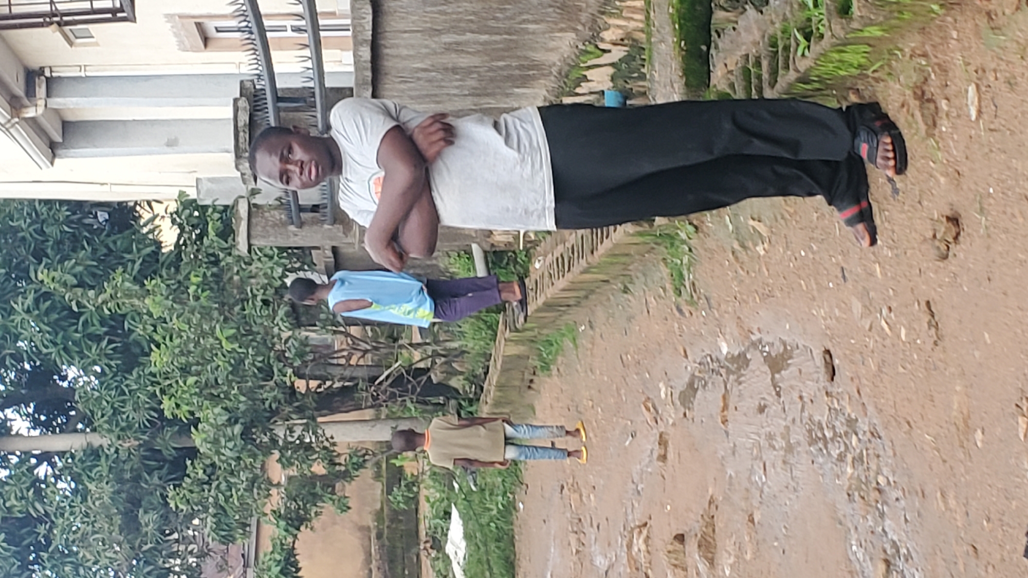 An image taken outdoors in Jos, Nigeria shows three young individuals standing on a dirt path. The individual in the foreground, a young male with dark skin and short hair, is facing the camera with his arms crossed. He is wearing a light grey t-shirt with a logo on the left chest and black pants, with black and red striped sandals. Behind him and to the left, another young male, facing away from the camera, is wearing a light green t-shirt and blue jeans with yellow sandals. Further back and to the left, a third young person, facing away, is wearing a light blue sleeveless top and dark purple pants. The background features lush green trees, a building with white walls and decorative metal fencing, and a low, moss-covered concrete wall. The ground appears to be a mix of dirt, stones, and puddles of water, suggesting recent rain.