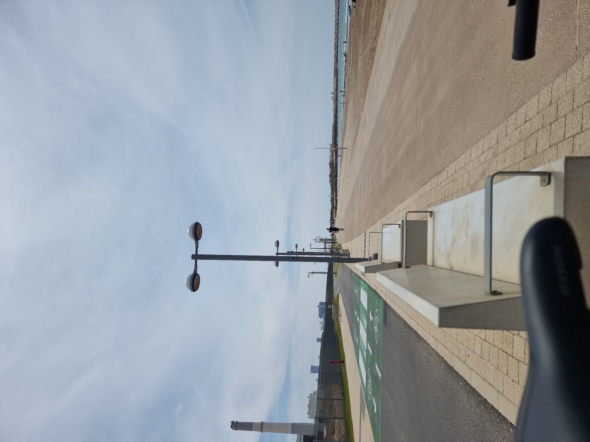 The image shows a bike path in Tel Aviv, Israel, next to the beach. The view is slightly skewed, taken from a moving bicycle, as indicated by the partially visible seat in the foreground. A green bike lane with white bicycle symbols runs along the path, bordered by a low retaining wall made of light-colored bricks. Adjacent to the bike path is a wide, sandy-colored walkway. In the distance, the Mediterranean Sea is visible. A series of streetlights line the bike path and walkway. Beyond the path, a fence separates it from a grassy area and buildings, including a tall smokestack. The sky is light blue with scattered clouds. Several people can be seen walking or cycling in the distance along the beach promenade. Bike racks are situated adjacent to the bike path.