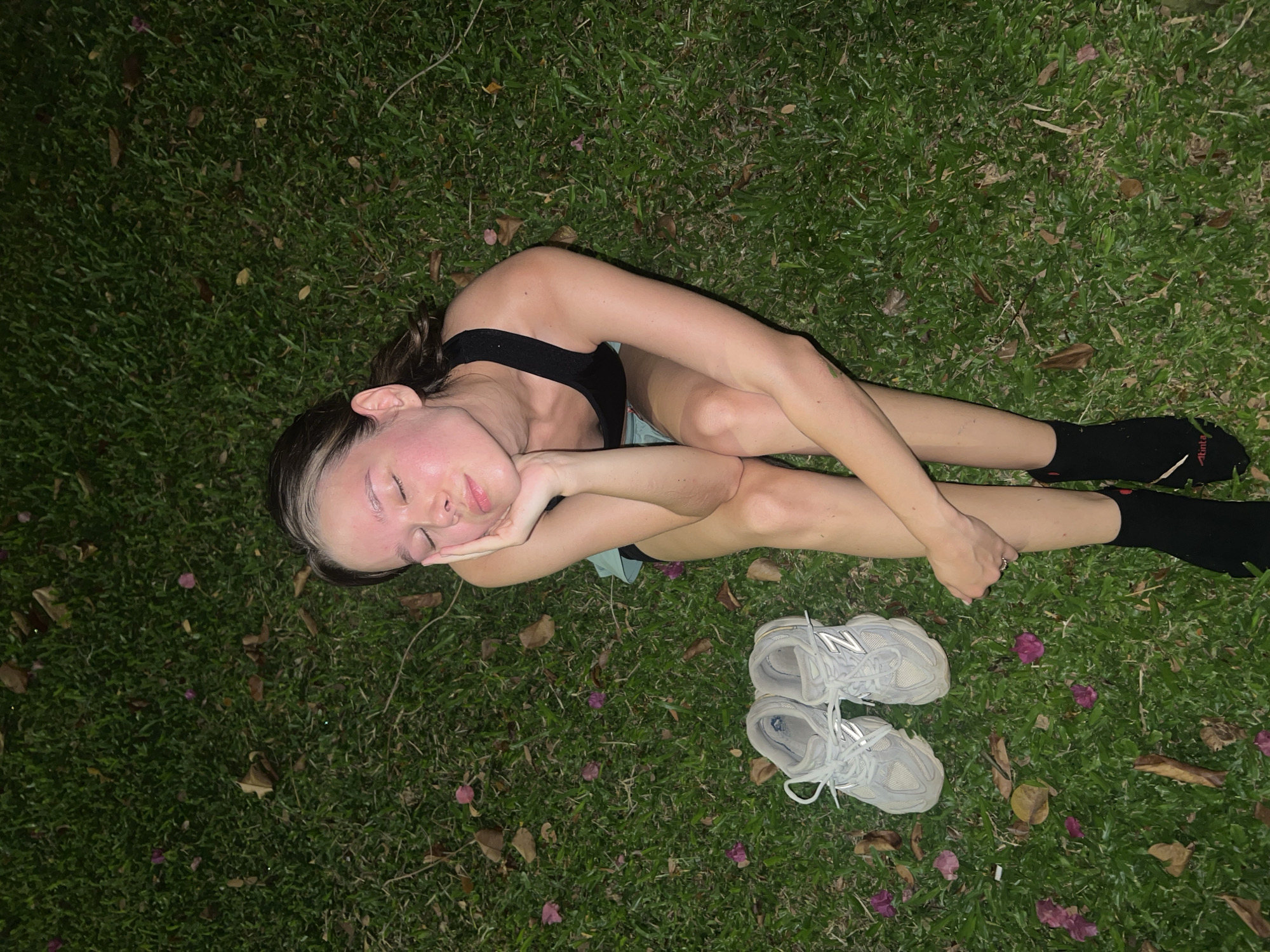 A young individual is captured in an overhead shot, lying peacefully on their side on vibrant green grass, likely in Nam Nha Trang, Vietnam. The person, appearing to be female, has dark hair with a distinctive white or blonde streak at the front, and her eyes are closed, suggesting rest or sleep. Her head is gently supported by her hands, which are crossed beneath her cheek. She wears a black athletic top or sports bra and light blue shorts, partially visible. Black ankle socks are on her feet; one sock features a red logo with white text that appears to read "Atleta."
Beside her feet, a pair of light grey New Balance sneakers with visible "N" logos are placed neatly on the grass. The ground is densely covered in green blades, interspersed with scattered brown leaves and delicate pink or purple flower petals, indicating an outdoor, natural setting. The lighting is dim and artificial, casting soft shadows, which points to the scene taking place at nighttime or late evening. The overall atmosphere conveyed is one of tranquility and relaxation.