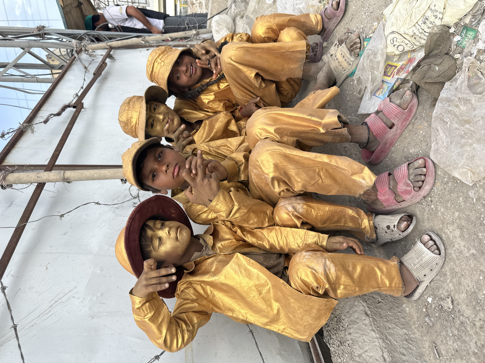 Four children painted gold from head to toe are seated on the ground near a low white wall with a metal structure and a man visible on top. They are wearing gold clothes and hats, and various sandals and the ground is littered with trash. The text indicates this is Quetta City Tehsil, Pakistan.