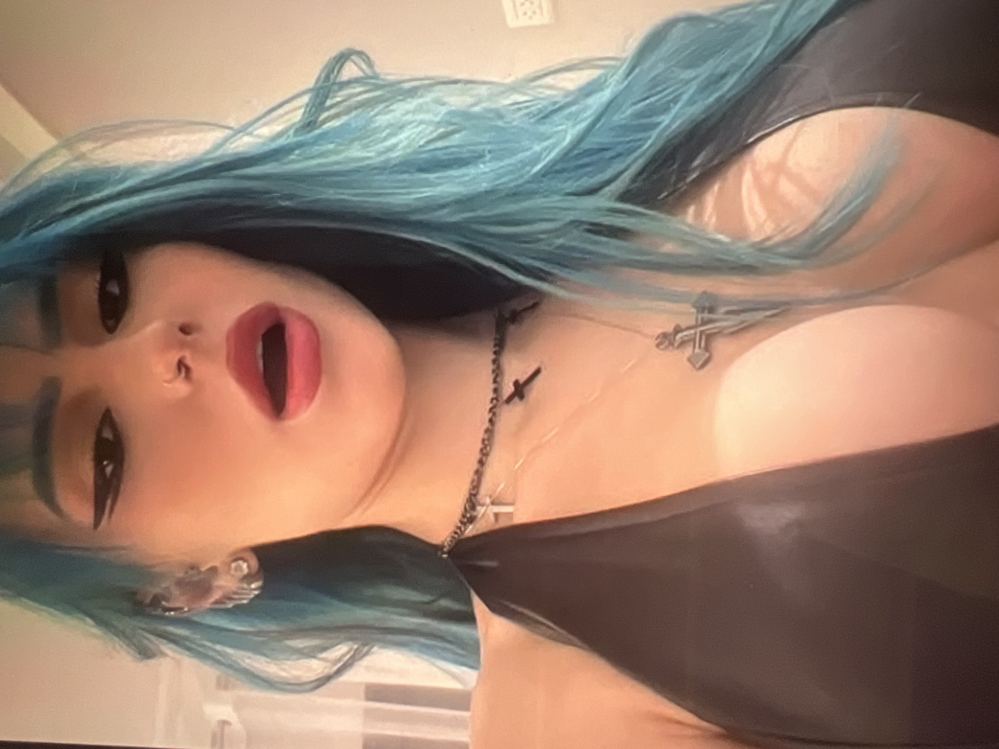 The image shows a close-up, slightly upward-angled, indoor shot of a person with teal-colored hair, light skin, and dark eye makeup. The person is wearing a black, low-cut top and a multi-layered necklace with cross pendants. Their mouth is open, and their head is tilted slightly. The background is out of focus and appears to be a wall with a light fixture. There is no identifiable information suggesting the location is Minsk, Belarus.