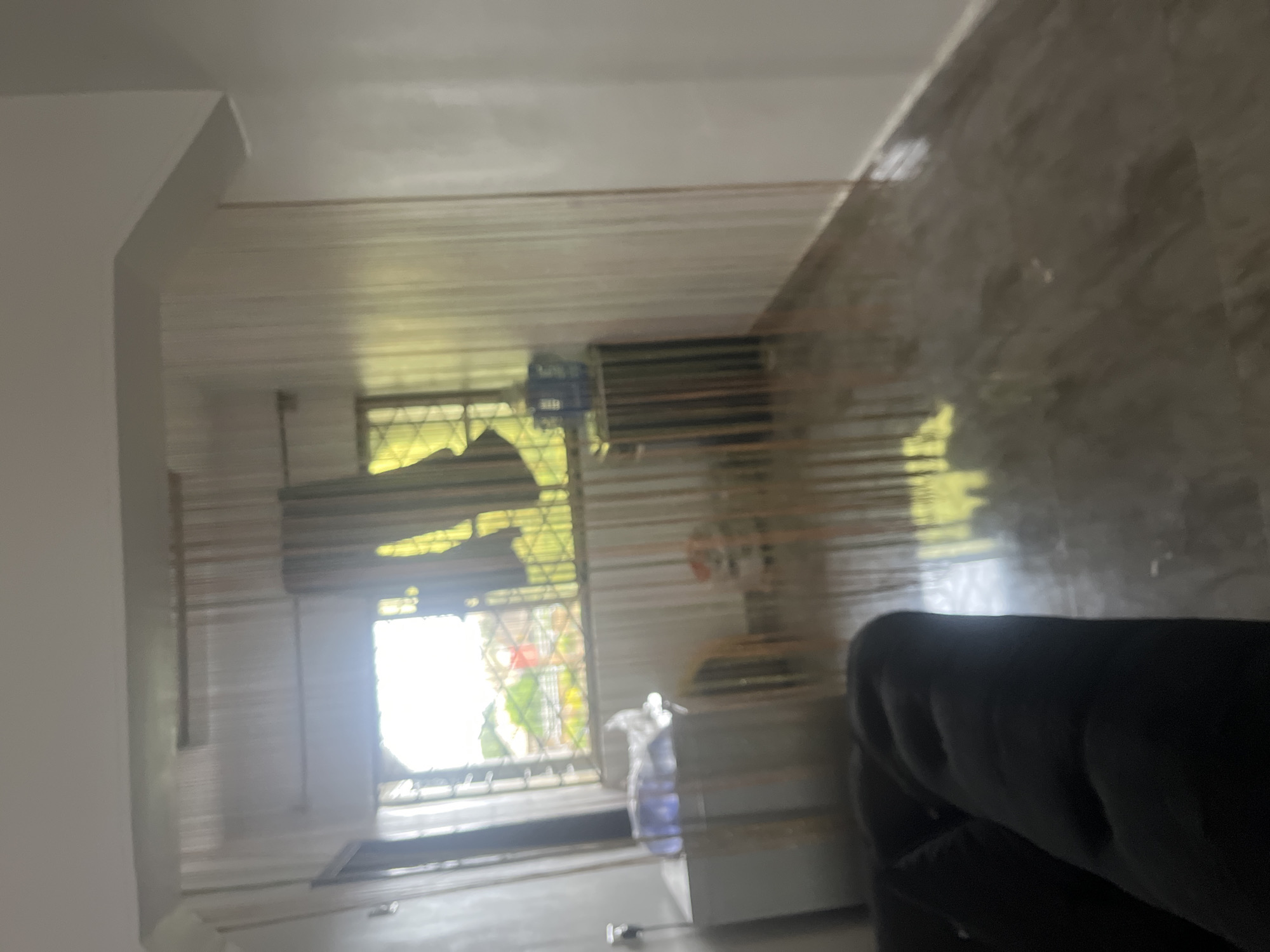 The image shows an indoor scene, possibly a room in a residential building in Jos, Nigeria. The room has plain walls and a tiled floor with a reflective surface. Light is coming in from a window, creating bright reflections on the floor. The window has a criss-cross grill and appears to be covered by blinds or curtains. Outside the window, greenery is visible, suggesting it is daytime.
There are several objects in the room, although many are indistinct due to the image's blurriness and reflections. One prominent object is a dark, plush sofa or chair in the foreground. In the middle ground, a blue object, possibly an air conditioning unit, is mounted on the wall near the window. There's also a clear plastic bottle with a blue liquid, possibly water, placed on a surface. The reflections on the floor suggest a busy or possibly a newly cleaned area.
The lighting indicates it is daytime, with bright sunlight streaming through the window. The overall impression of the scene is one of a lived-in space, perhaps a living room or lounge. There is no visible text in the image. The low resolution and blurriness obscure finer details and prevent identification of specific activities or emotions.
