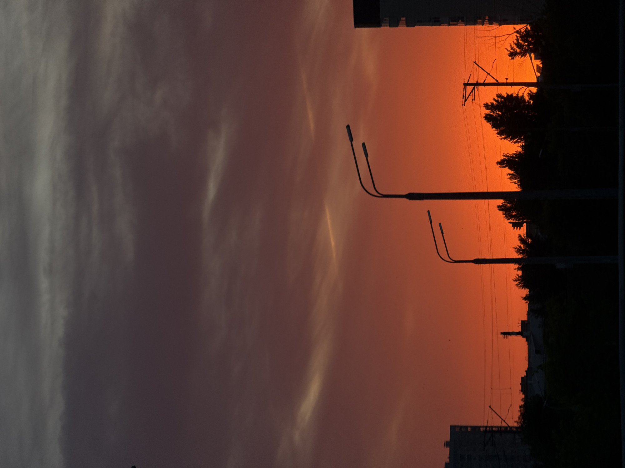 The image captures a sunset in Kazan, Russia. The sky transitions from a dusky grey with visible cloud formations on the left to a vibrant orange on the right. Two street lamps are silhouetted against the orange sky. In the distance, there are outlines of buildings and trees, including power lines.