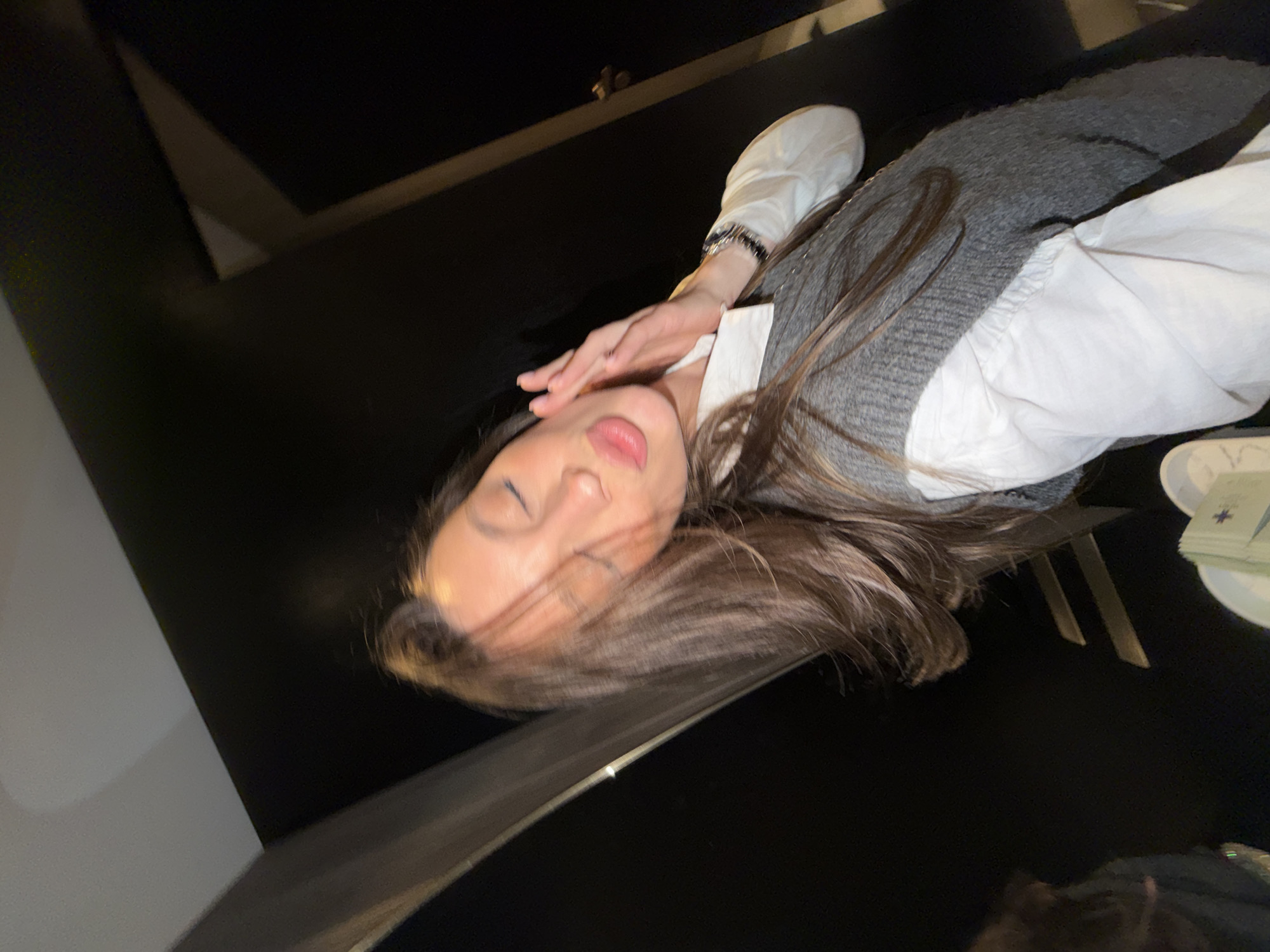 A young woman is lying on a dark, glossy surface, likely a table, at a dining establishment in Budva, Montenegro. She is wearing a white collared shirt with a gray sweater vest over it. Her long brown hair is spread across the table. Her hand rests near her face. A stack of napkins and a small plate are visible near the edge of the table, suggesting this is a restaurant or bar. The background is dark and out of focus.