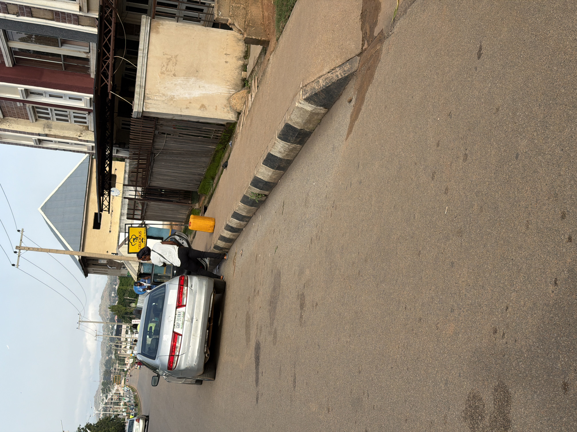 In Gura Top, Nigeria, a silver sedan is parked on the side of a paved road, angled towards the sidewalk. A person wearing a white top and black pants stands beside the open driver's side door, facing away from the camera. Adjacent to the car, on the sidewalk, is a bright yellow plastic container. The street has a distinctive black and white striped curb. In the background, a multi-story building with a reddish-brown facade and numerous windows stands next to a wooden fence. Further down the street, hills and distant trees are visible under a cloudy sky.