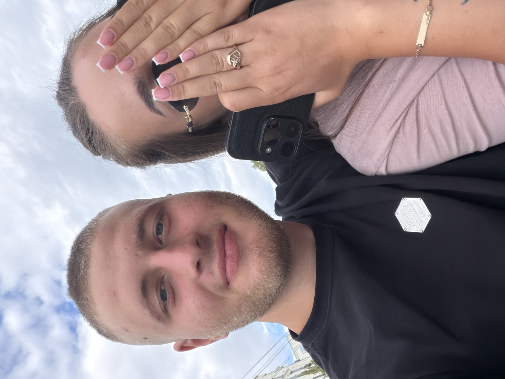 A young man with a buzz cut and light stubble smiles at the camera, his head and shoulders filling the left side of the frame. He is wearing a black t-shirt with a hexagonal silver logo on the left chest. To his right, a young woman is partially visible, holding a black smartphone in her right hand and covering the lower half of her face with her left hand. She has long brown hair pulled back, and is wearing sunglasses. Her visible hand displays a gold ring with a stylized "M" shaped design, and her fingernails are painted with white tips. She is wearing a light pink t-shirt. A gold bracelet with heart charms is visible on her left wrist. The background shows a cloudy sky and a building facade to the left. The overall scene appears to be an outdoor selfie taken in the city of Strășeni, Moldova, judging by the building architecture and the visible sky.