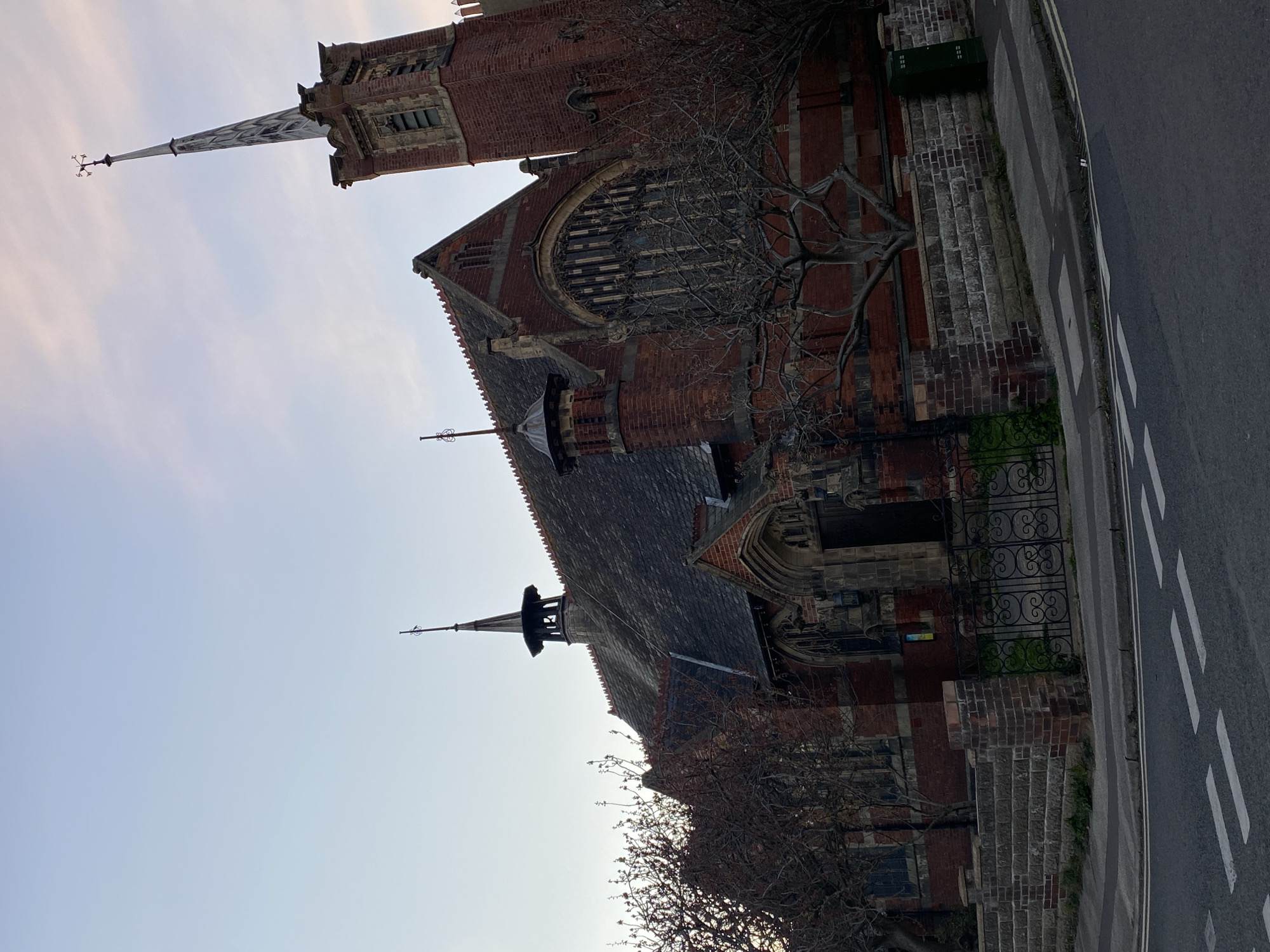 The image captures an architectural structure, primarily constructed of red brick. A prominent steeple rises high, topped with a weather vane. An arched window is a focal point of the building, partially obscured by bare tree branches. The building sits adjacent to a sidewalk and road, marked with white stripes. The sky above is pale and light. This is Saint John's church in Felixstowe, East Suffolk, United Kingdom.