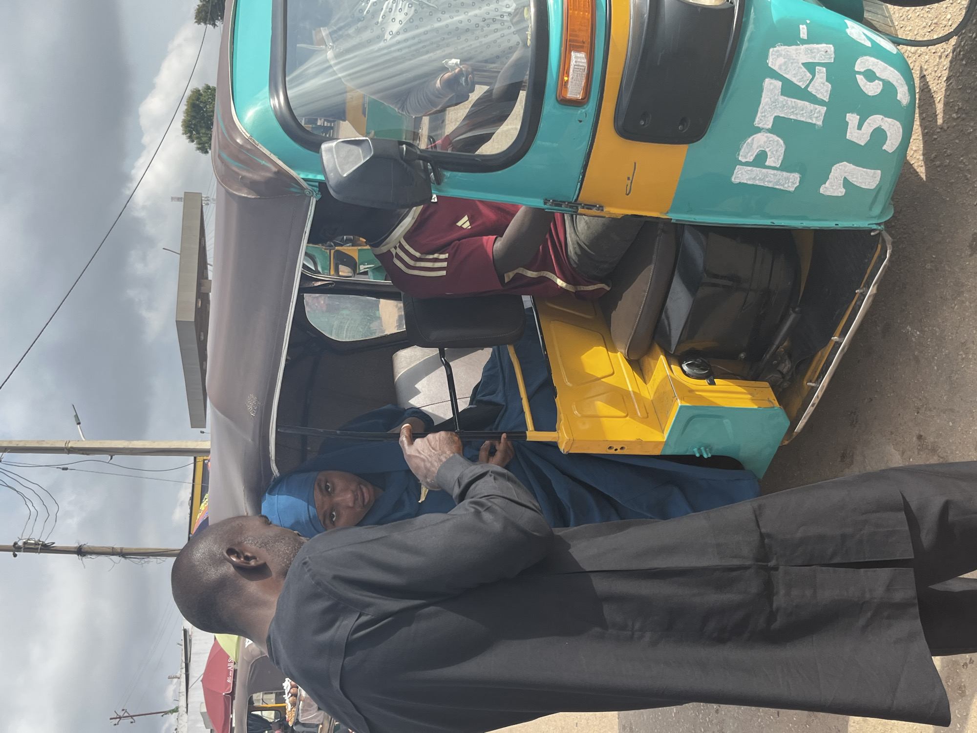 In Gura Top, Nigeria, a man in a black traditional garment engages in a conversation with a woman wearing a blue hijab inside a teal and yellow auto-rickshaw. The auto-rickshaw has "PTA-759" written in white on its side. The driver, wearing a maroon and white striped shirt, is seated in the driver's seat. The background shows a cloudy sky, utility poles, and what appears to be a gas station. The scene suggests a street-level interaction, possibly a transaction or negotiation, involving transportation.