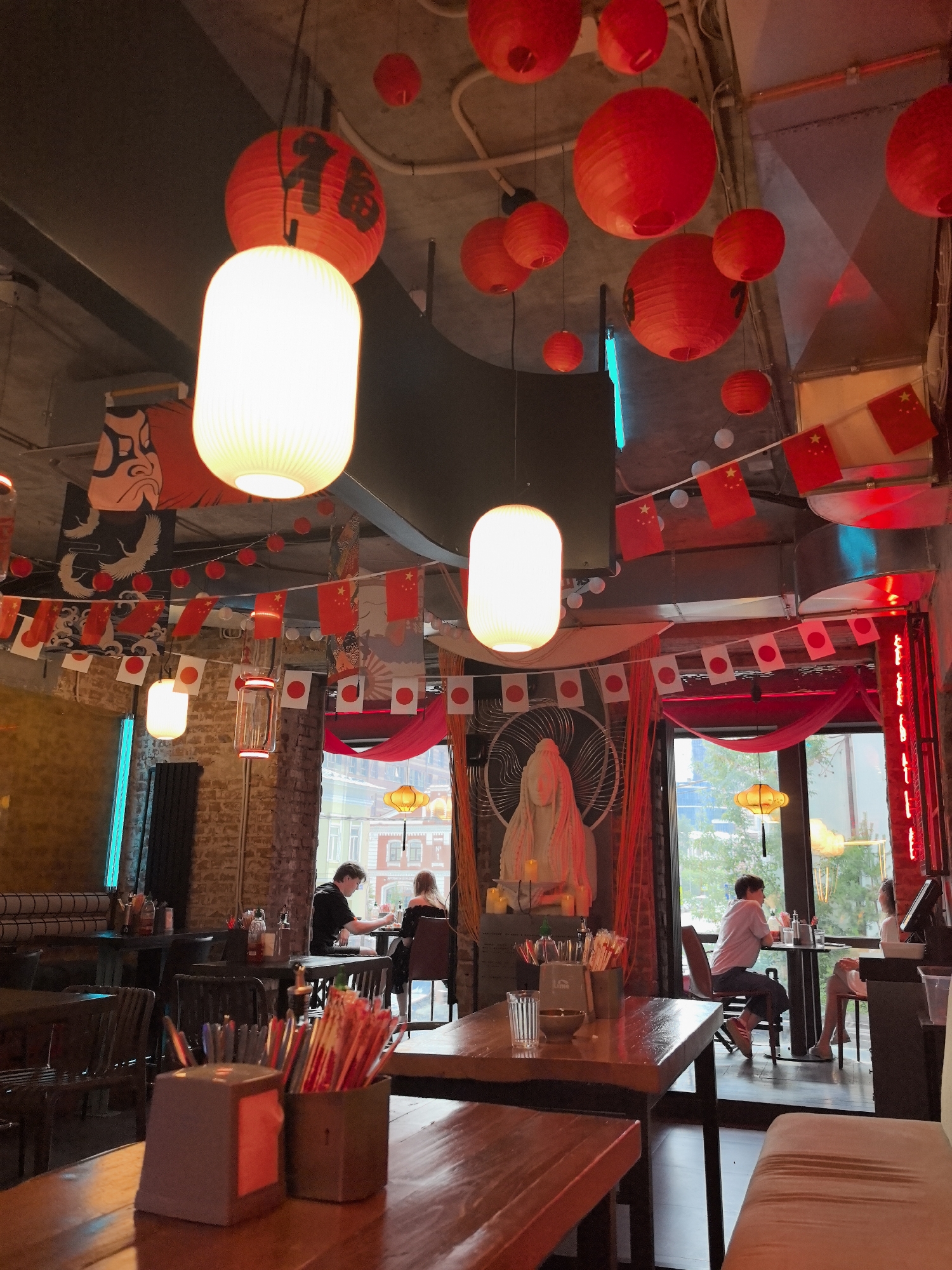 This image depicts the interior of an Asian-themed restaurant, possibly in the city of Perm, Russia, given the presence of Russian Cyrillic text on a sign in the upper left quadrant. The restaurant is decorated with numerous red paper lanterns hanging from the ceiling, some adorned with black calligraphy. Bunting flags of Japan and China are also displayed, creating a festive atmosphere. The walls are a mix of exposed brick and dark paneling.
Several tables with wooden tops and chairs are visible, some occupied by patrons. In the foreground, a wooden table holds containers of chopsticks and pens. In the middle ground, two individuals are seated at a table near a large window overlooking an outdoor scene. One person is facing away from the camera, while the other is partially visible. Further back, more tables and seating arrangements are present. The lighting is dim, with the red lanterns and a bright blue neon light providing illumination. A large, stylized statue or artwork is visible on the left side of the background, behind a red curtain. The overall impression is that of a bustling, possibly themed, dining establishment.