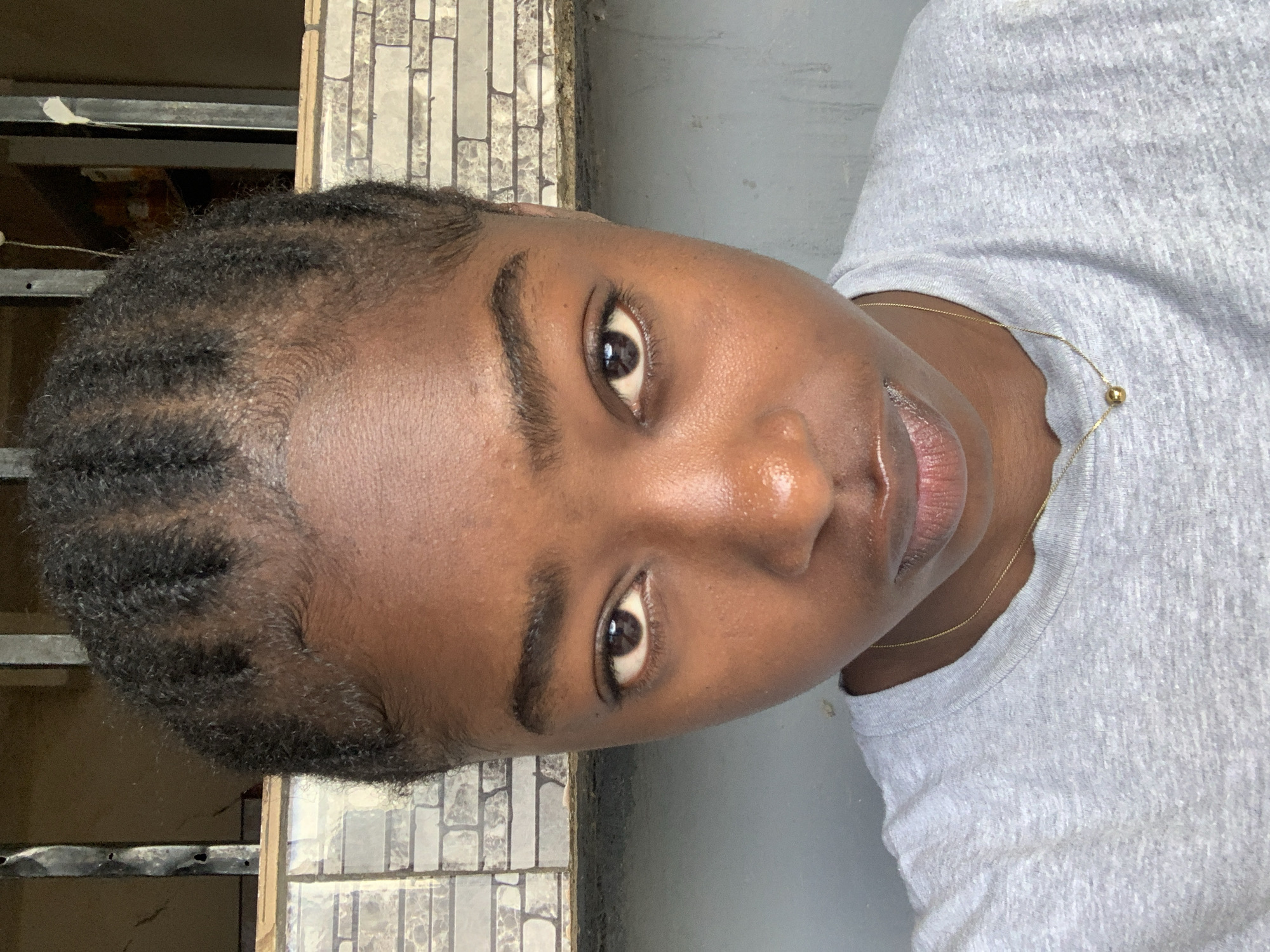 A medium, close-up angled shot features a dark-skinned person in a light grey shirt. The person has short, dark hair styled in thin braids. The person's eyes are open and looking toward the camera. The person has dark eyebrows and dark pupils. A thin gold chain necklace is visible around the person's neck. The background includes tiled walls of varied colors, grey concrete, and a metal and wood structure. The image was likely taken in Kabong, Nigeria.