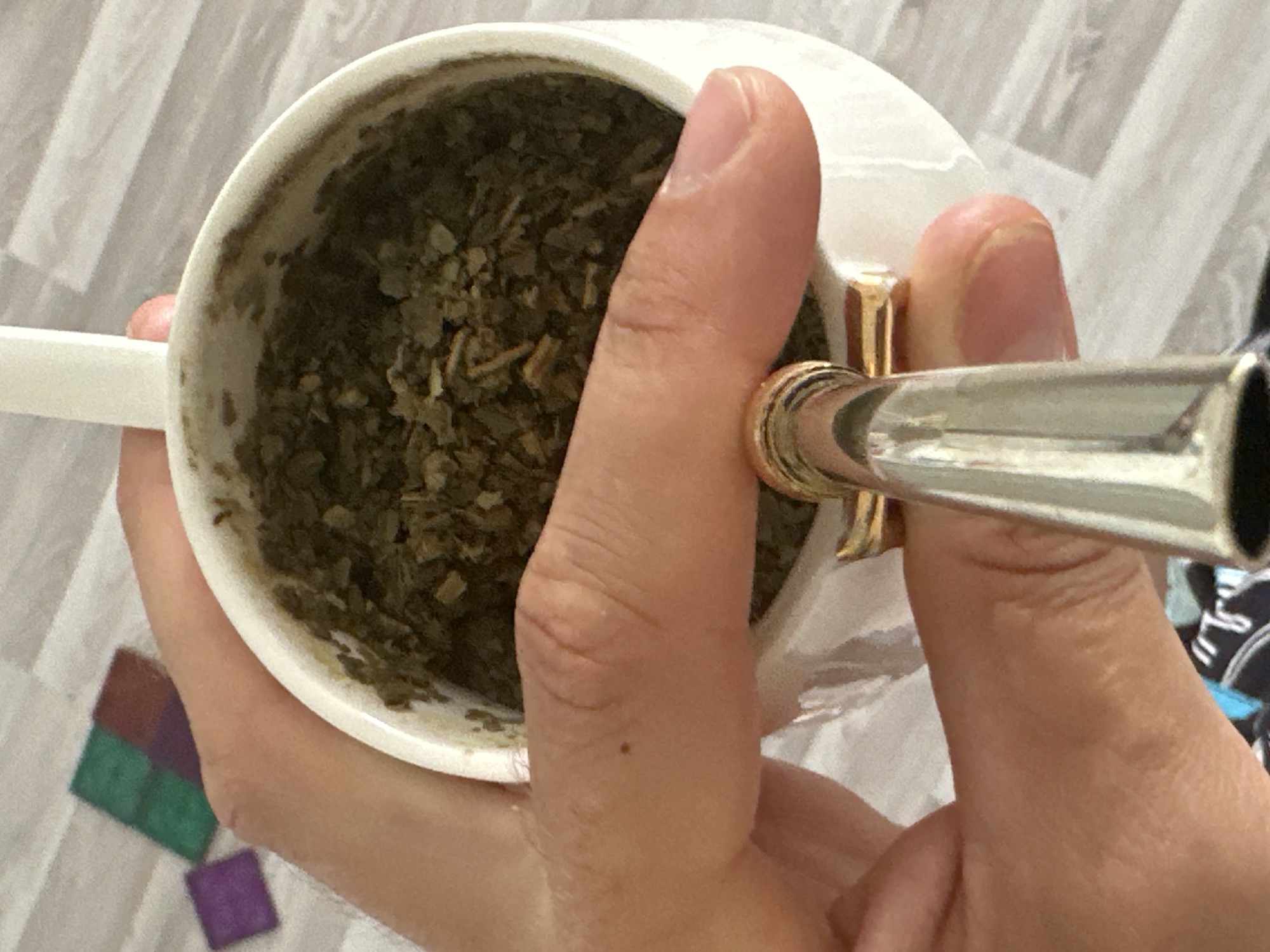 A close-up, high-angle shot shows a person's hands holding a white ceramic cup, likely a mate gourd, filled with dried yerba mate leaves. A silver-colored metal bombilla (straw with a filter) featuring a gold-colored band is inserted into the mate. The person's thumb is prominently visible, resting against the cup, while other fingers cradle the vessel. The activity depicted is the holding and implied consumption or preparation of yerba mate.
The background consists of a light-colored, textured surface, possibly a patterned blanket or flooring, with some slight wrinkles visible. In the bottom left corner, several small, colored square objects—green, red-brown, and purple—are scattered on the surface. Another part of the background shows a dark blue fabric with white patterns, possibly part of the person's clothing or another item nearby. The setting appears to be indoors, under ambient lighting, in Fethiye, Turkey. No text is visible in the image.