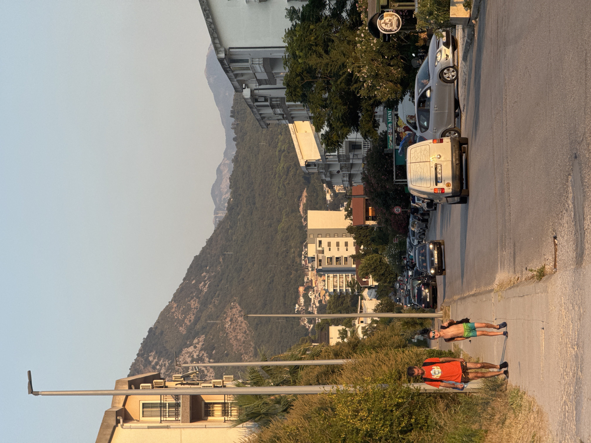 A street scene in Sutomore, Montenegro, features two males walking on a sidewalk in the foreground. The male on the left, taller and wearing an orange t-shirt and dark shorts, carries a backpack. The shorter male on the right wears swim trunks and sunglasses, also carrying a backpack. The sidewalk is paved with concrete. Behind them, a road is lined with parked and moving vehicles, including a white van and a silver hatchback. Buildings with balconies and air conditioning units are visible along the street. In the background, a densely populated town is nestled against a lush, green, mountainous terrain under a clear sky. The sun casts a warm light on the scene, indicating it is either morning or late afternoon.