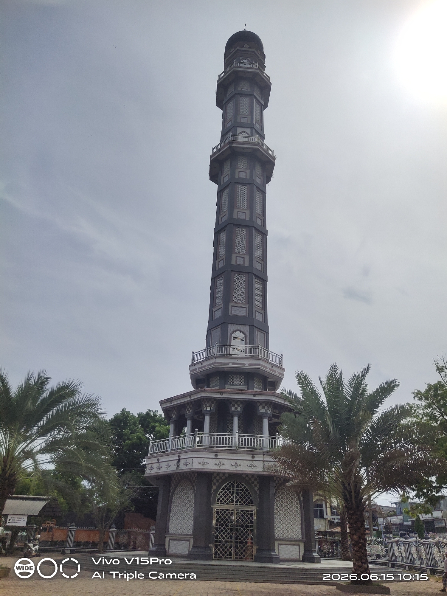 The image shows a tall, dark gray minaret with an octagonal base, topped with a dome. The minaret has multiple levels with balconies and decorative windows. Palm trees are visible in the foreground, to the left and right of the minaret. The sky is overcast with some clouds. A timestamp in the lower right corner reads "2026.06.15 10:15". A logo and text from "Vivo V15Pro AI Triple Camera" are present in the lower left corner. In the background, there are some buildings and other trees. The scene appears to be outdoors in a public space. Based on the metadata and the name of the minaret, the location is identified as Keudekruenggeukueh, Indonesia.