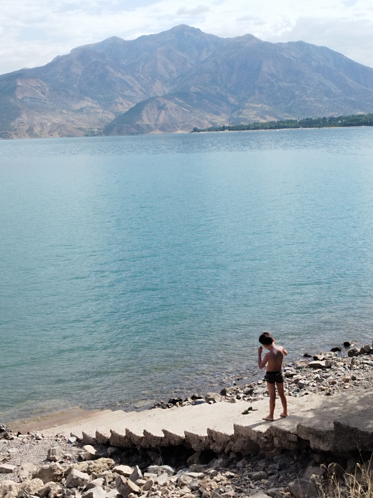 In a wide shot, a young boy stands on a rocky shoreline facing a vast expanse of tranquil, turquoise water. The water's surface is slightly rippled under a hazy, overcast sky, and in the distance, a range of imposing, layered mountains dominates the horizon. The boy, wearing dark patterned swim trunks, is positioned on a concrete embankment dotted with rocks and debris. He is looking down at his hand, which is raised to his face, as if picking something off or examining it. The shoreline is a mix of pebbles, larger stones, and broken concrete structures, creating a rugged, textured foreground. The overall scene evokes a sense of quiet solitude in a natural, mountainous landscape.
