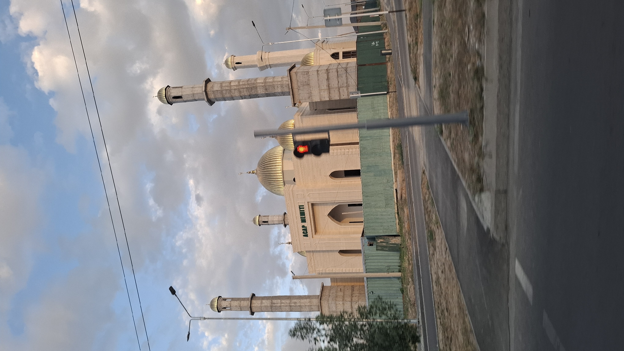The image shows a mosque with golden domes and minarets, likely the "Asar Memiti" mosque in Shymkent, Kazakhstan. The mosque is constructed of light-colored stone and features traditional Islamic architectural elements. In front of the mosque, there is a green fence and a road with a traffic light that is red. The sky is cloudy with patches of blue. A street lamp and some trees are also visible in the foreground, suggesting an urban or suburban setting. The mosque appears to be under construction or recently completed, with some unfinished elements visible on the minarets.