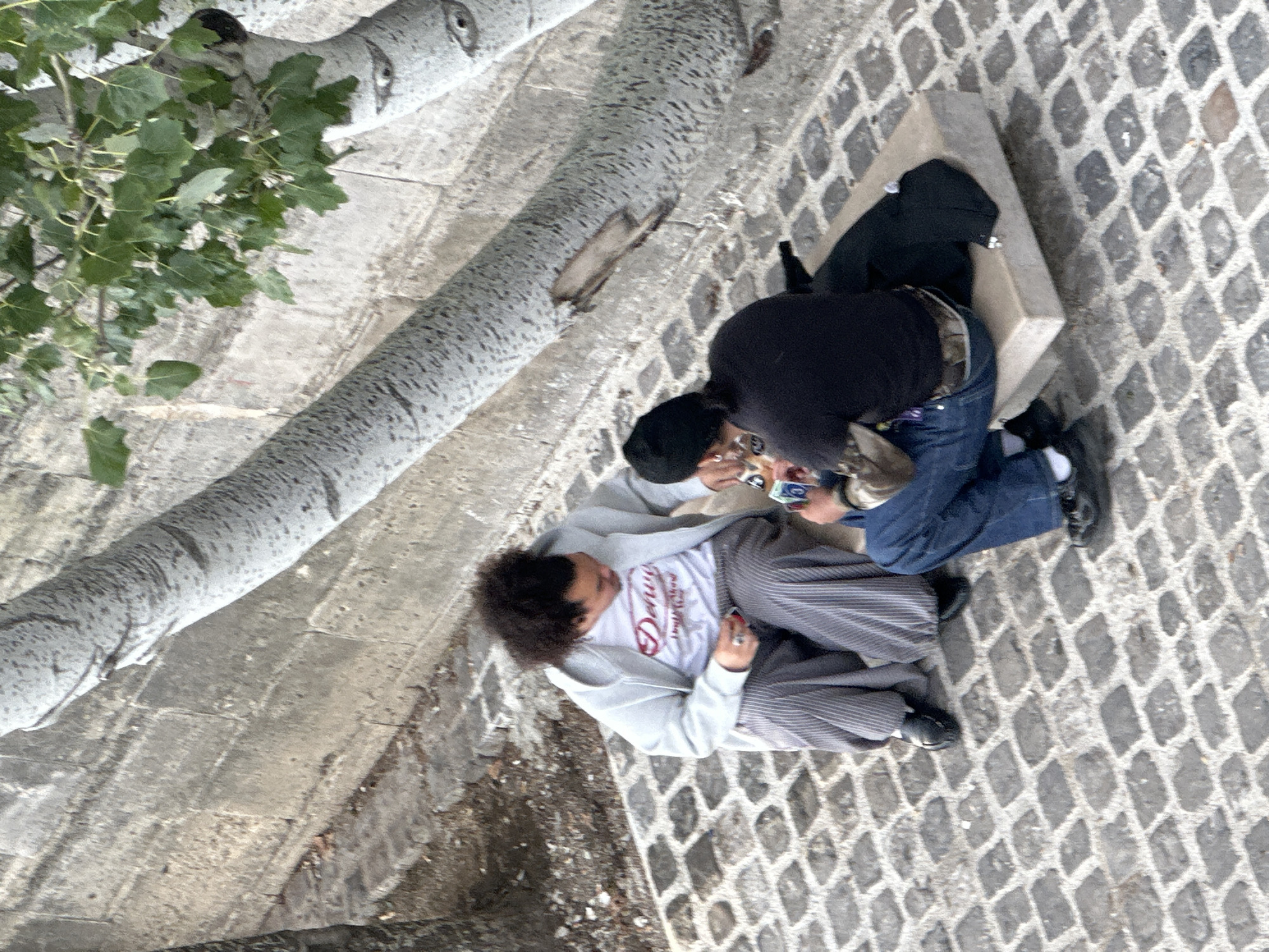 Two people are sitting on a stone bench in an outdoor setting. One individual, with curly dark hair, is wearing a light gray hoodie over a white t-shirt with a design on it, along with striped gray and white pants. They are looking down at something in their hands. The other individual, wearing a black top and jeans, is also looking down. There is a black bag or jacket on the bench beside them. The ground is paved with cobblestones, and a tree branch with green leaves is visible in the upper-left corner. The bench and the people are near a stone wall or embankment. Given the architectural style and the type of pavement, the location could be in Paris, France. The two people appear to be looking at something together.
