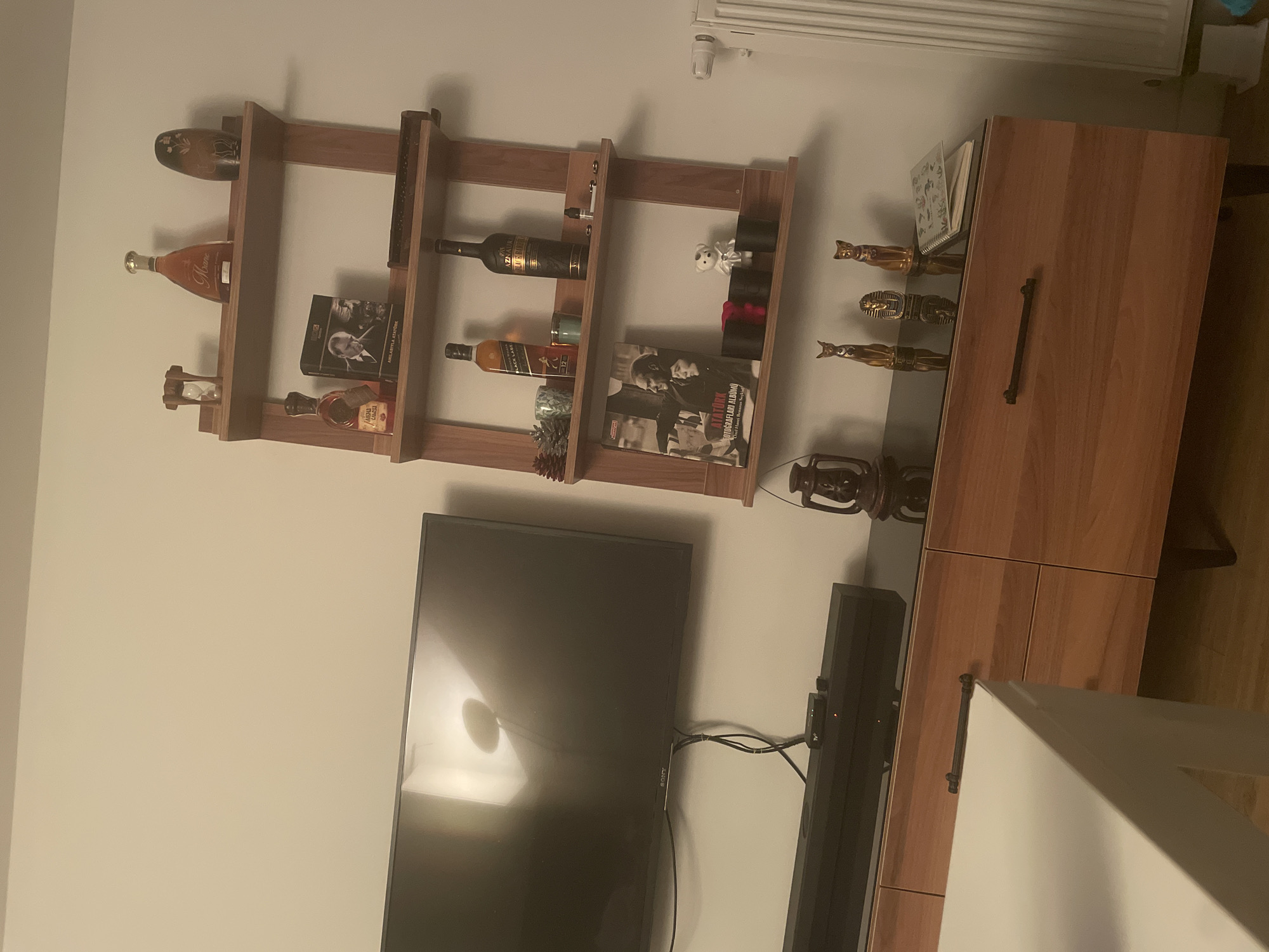 The image depicts an indoor scene, likely a living room, in Yunus Emre, Turkey. A large, dark-screen television is mounted on a light-colored wall to the left of the frame. Below the television, a black soundbar is positioned on top of a wooden media console with a dark glass top. The media console features several drawers with dark handles.
To the right of the television, a multi-tiered wooden shelf unit is mounted on the wall. The shelves are adorned with various decorative items, including bottles of alcohol, books, and small figurines. Notable items on the shelves include an hourglass, a bottle labeled "Mone," a dark brown decorative vase, a box with an image of Mustafa Kemal Atatürk, a bottle of Johnnie Walker Black Label, another dark bottle, a small lantern, and several cat figurines and an Egyptian pharaoh figurine on the media console. A white teddy bear and two black cylindrical containers are also visible on one of the shelves. A spiral-bound notebook rests on the media console.
The overall environment appears to be a residential space with a modern aesthetic, characterized by the combination of the wooden furniture and the minimalist wall-mounted shelves. The lighting is dim, suggesting an evening or indoor setting.