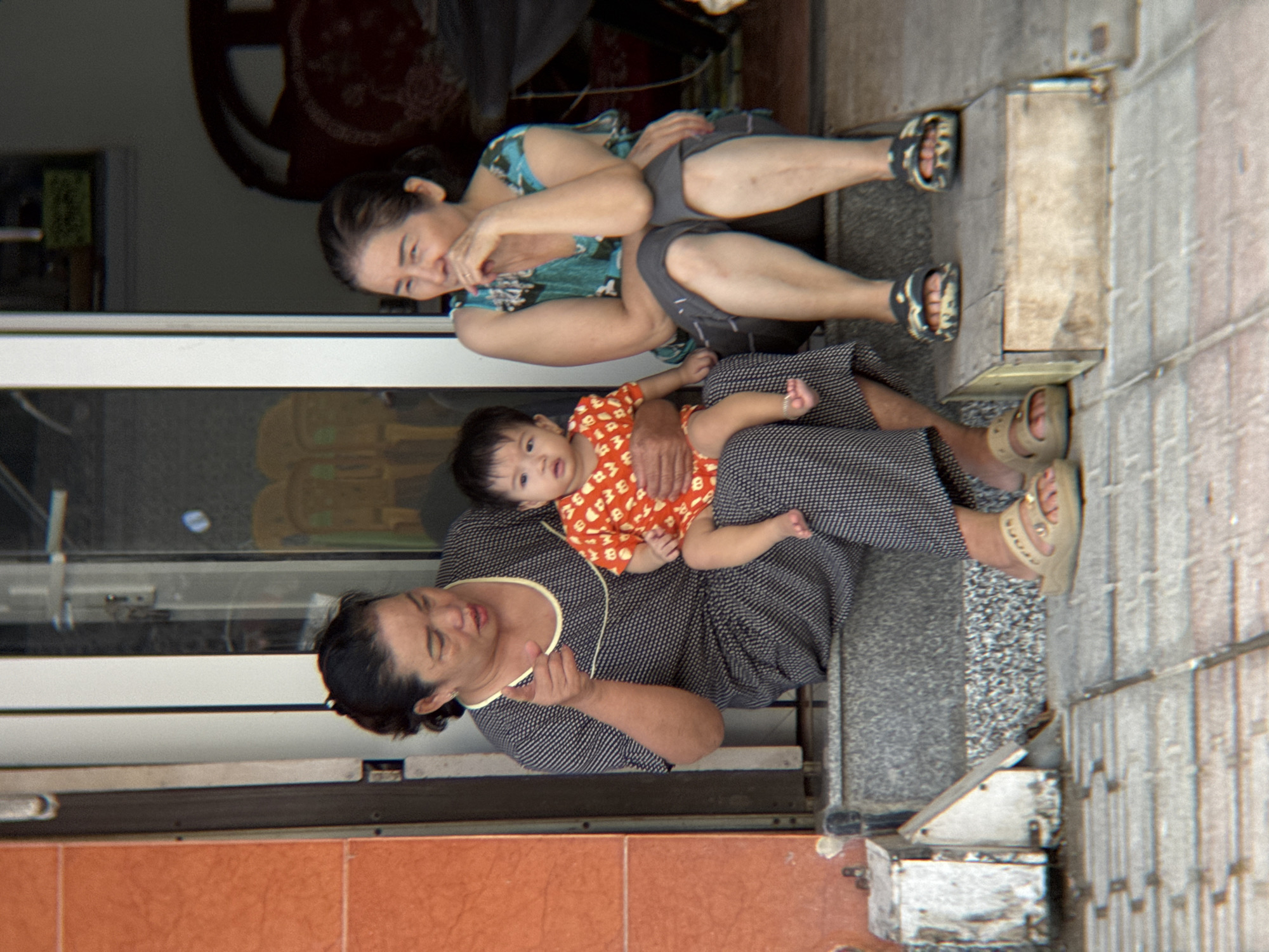 Two Vietnamese women and a baby are sitting on steps outside a building. The woman on the left is holding the baby on her lap. She is wearing a dark grey dress with white polka dots and yellow trim. The baby is wearing an orange and white patterned outfit. The woman on the right is wearing a blue and green patterned top and grey shorts. She is resting her chin on her hand. They are all wearing sandals. The building appears to be a shop or restaurant. The image is taken in Nha Trang, Vietnam.