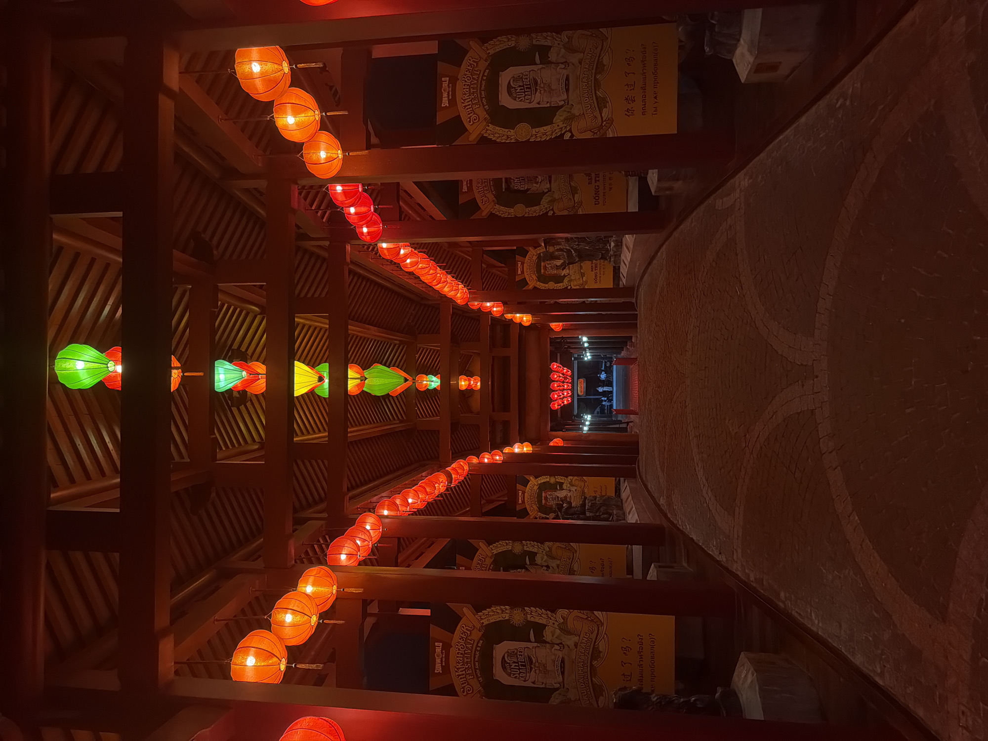 The image captures a long, illuminated walkway or corridor at night, featuring traditional Asian architecture and festive decorations. The scene is set within a covered structure, likely a part of a resort or cultural attraction in Đà Nẵng, Vietnam, such as Sun World Ba Na Hills.
The walkway is flanked by robust dark wooden pillars and beams, supporting a wooden ceiling from which numerous colorful lanterns are suspended. On the left side, the lanterns are primarily green and yellow, while the right side is adorned with long rows of red and orange lanterns, casting a warm glow. The pathway beneath is paved with bricks or stones, displaying subtle circular patterns.
Along the right side of the corridor, large vertical banners are prominently displayed, advertising a "Sunkka Beer Festival." These banners feature a cartoon-style graphic of a character holding a beer and text in multiple languages. Visible phrases include "SUNKRAFTBEER FESTIVAL," "LỄ HỘI ẨM THỰC & BIA" (Vietnamese for "Food & Beer Festival"), "UỐNG THỬ" (Vietnamese for "Try"), "YOU'RE INVITED!," and "LAU DAI HAT TRANG." Multilingual questions asking "Have you tried it?" are also present in Chinese ("你尝过了吗?"), Thai ("คุณลองชิมแล้วหรือยัง?"), and Russian ("Ты уже пробовал(а)?").
The time of day is clearly night or late evening, as indicated by the darkness and the lanterns being the main light source. The ambiance is inviting and festive, suggesting a cultural event or a popular tourist pathway. No individuals are visible in the frame.