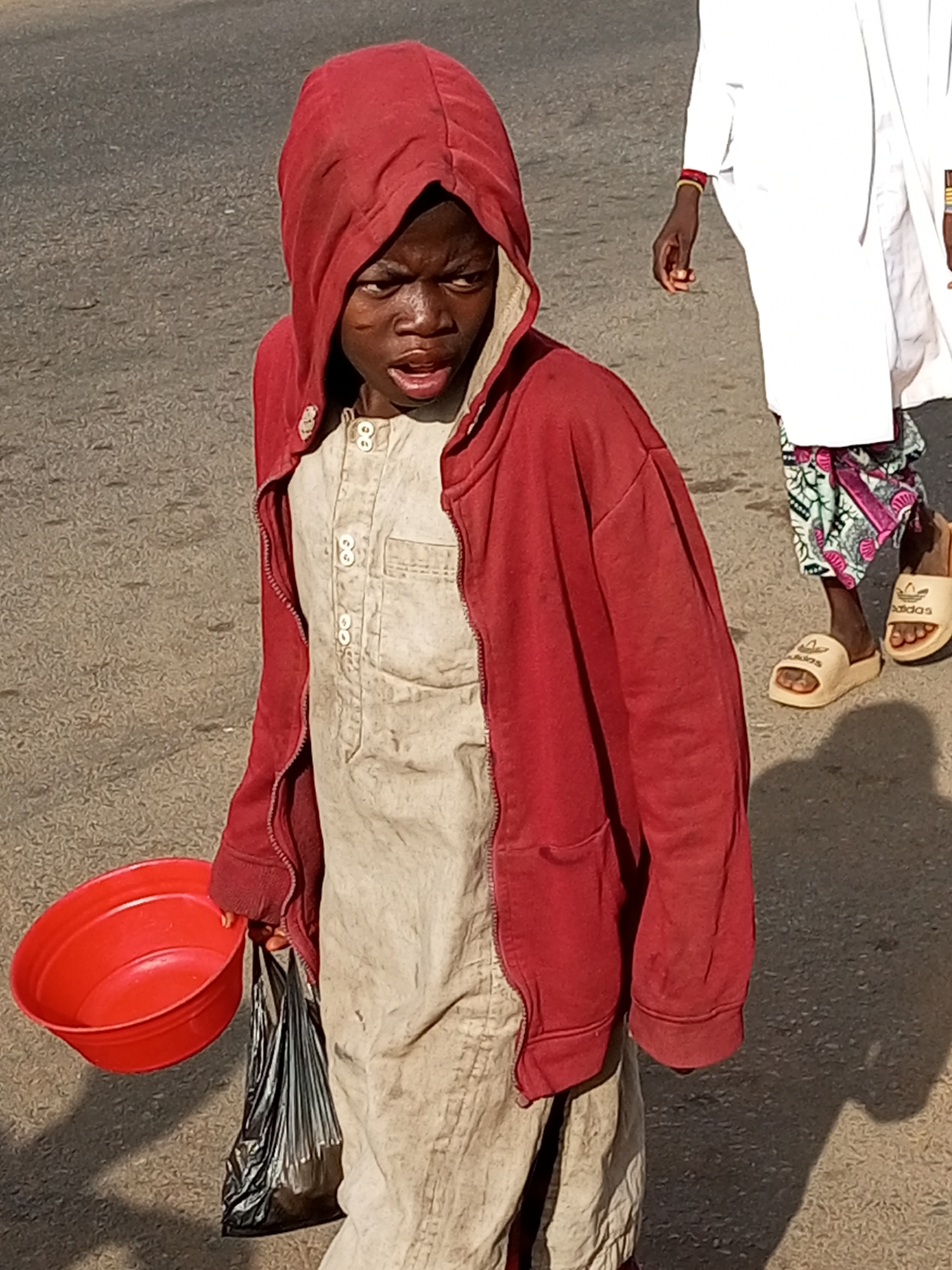 In Gombe, Nigeria, a young individual wearing a red hooded jacket over a light-colored tunic, potentially a traditional attire, stands on a paved surface. The individual's clothing appears worn and dusty. They are holding a bright red bowl in their left hand and a black plastic bag in their right. The individual's facial expression is earnest, with their mouth slightly open as if speaking or calling out. Behind the individual, another person is partially visible, wearing a white garment and patterned trousers, with sandals on their feet. The ground is asphalt, typical of a street or road in a city. Shadows from the individuals and potentially other unseen objects are cast on the ground, indicating daylight conditions. The scene suggests a street-based interaction, possibly related to commerce or begging.