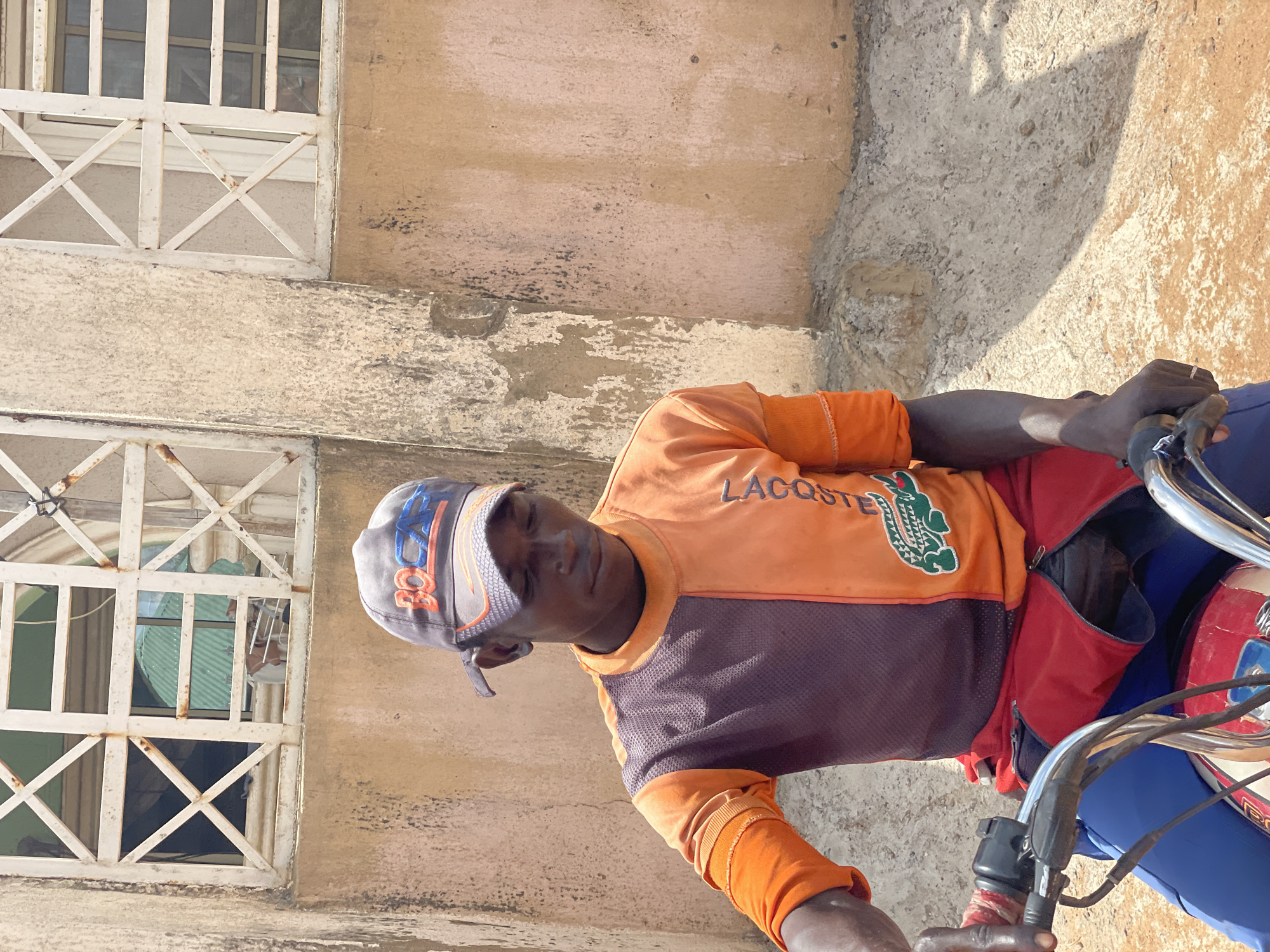A man on a motorcycle sits next to a building. The man is wearing an orange and purple Lacoste shirt and a baseball cap. The motorcycle is blue and red. The building has two windows with white frames and an off-white facade. The image was taken in daylight, and the location may be Lokoja, Nigeria.