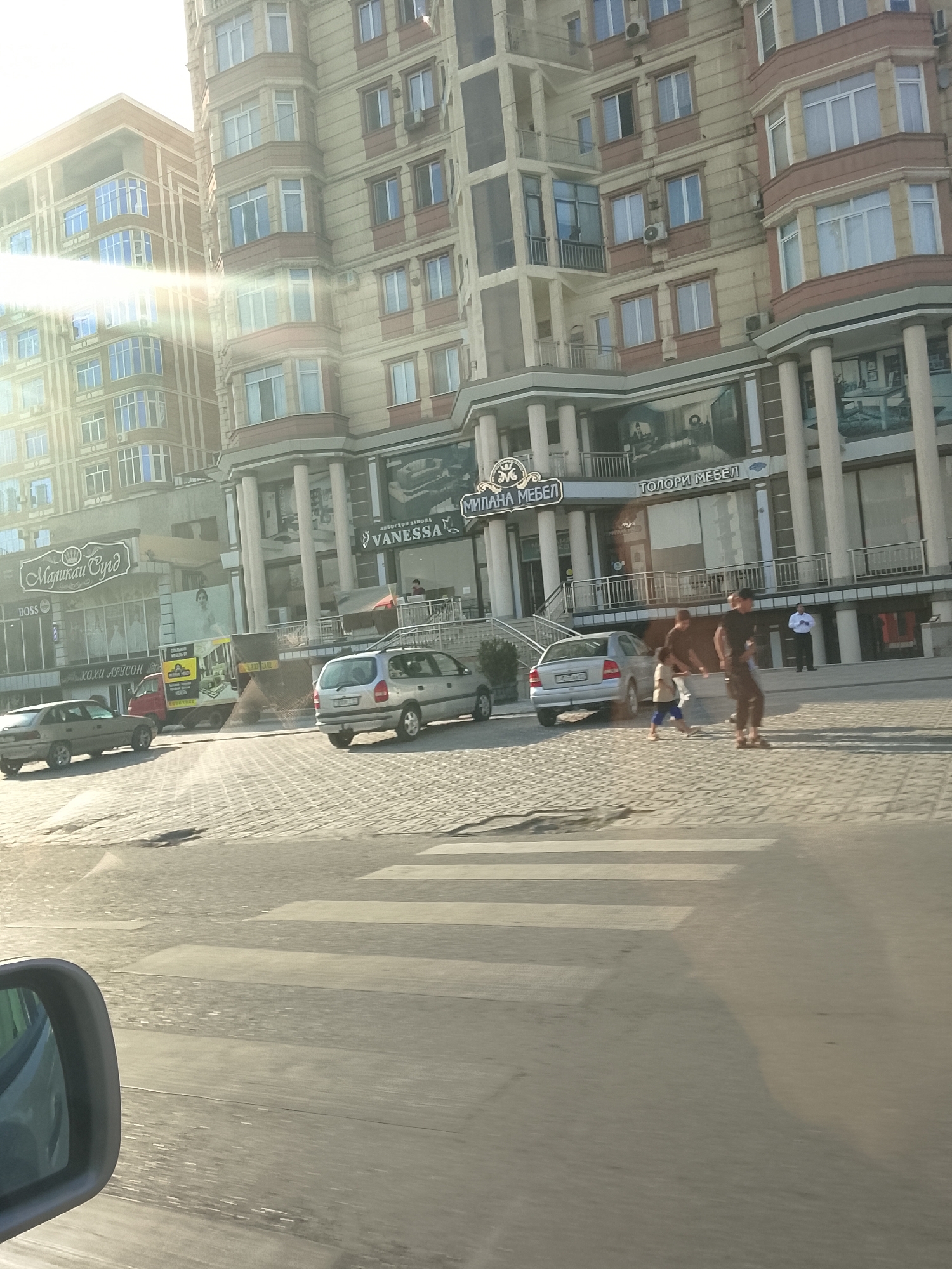In Khujand, Tajikistan, a sun-drenched street scene unfolds. A multi-story building with beige and brown facades dominates the background, featuring numerous windows and balconies, some adorned with decorative elements. The building houses various businesses, including "BOSS," "Маликаи Суд," "VANESSA," "МИЛАНА МЕБЕЛ," and "ГОЛОРИ МЕБЕЛ," indicated by signage. A red truck with advertising is parked nearby. On the street, a crosswalk is visible. Several cars are present: a silver sedan is parked on the left, a silver hatchback is driving on the crosswalk, and a silver station wagon is parked on the right. Three individuals are walking across the crosswalk: a man in a dark t-shirt and pants, a child in a light-colored shirt and blue pants, and another man in a dark t-shirt and dark pants. A person in a light blue shirt stands near a building entrance. The lighting suggests it is either early morning or late afternoon due to the long shadows and the bright sun glare. A side-view mirror of a vehicle is in the foreground on the left.