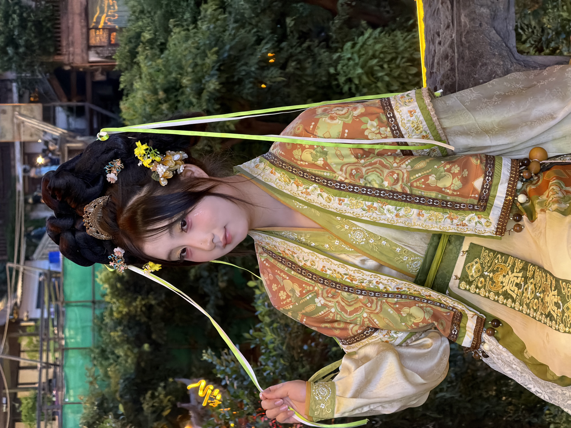 In Yuzhong District, China, a young East Asian woman, with dark hair elaborately styled into an updo adorned with flowers and hairpins, is pictured outdoors at dusk. She is wearing traditional Chinese attire, a pale gold silk garment layered with an ornate outer robe featuring intricate patterns of flowers, fruit, and geometric designs in shades of green, orange, and brown. The outfit includes a sash and decorative elements like beads. She is looking towards the camera with a slight smile, holding a green and white ribbon from her hair. The background consists of lush green foliage, with blurred lights and structures suggesting an urban or semi-urban setting at night.