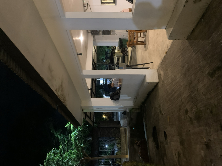 The scene is an outdoor area at night in Probolinggo, Indonesia, at coordinates -7.751 latitude and 113.213 longitude. The view is from a covered, concrete patio. Two people are visible, one sitting and looking at a laptop screen, the other sitting at a table. The patio is illuminated by artificial light. Behind the patio are two cars parked under a covered area. There is a large tree on the left side of the patio and a concrete wall to the right. A wooden table and chairs are situated in the center of the patio with a trash can behind the chairs. The patio is surrounded by a white concrete wall. There are several lights on the patio and a small, illuminated sign on the wall.