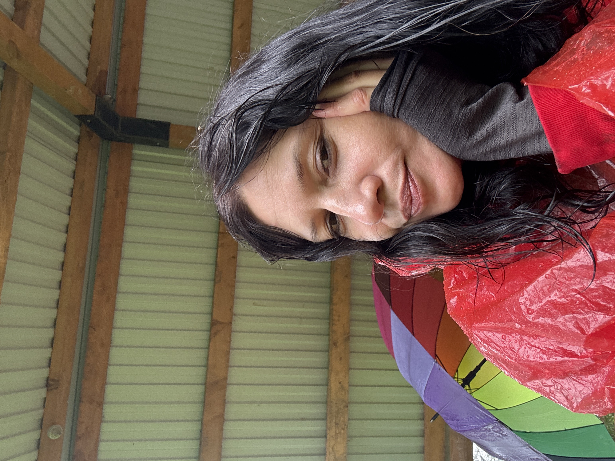 A woman with long, dark, wet hair and light brown skin is leaning her head on her hand, resting her cheek on her knuckles. She is wearing a red, translucent, crinkled raincoat with water droplets on it. Underneath, she is wearing a dark gray long-sleeved shirt. To her left, a rainbow-colored umbrella is visible, with segments of purple, blue, green, and orange. The background consists of a light green corrugated metal roof supported by wooden beams. The scene appears to be outdoors, possibly under a covered structure like a shed or awning, given the visible roof and beams. The overall impression is one of a person seeking shelter from rain. There is no discernible information in the image that would indicate the location is in the city of Nagornskiy, Russia.