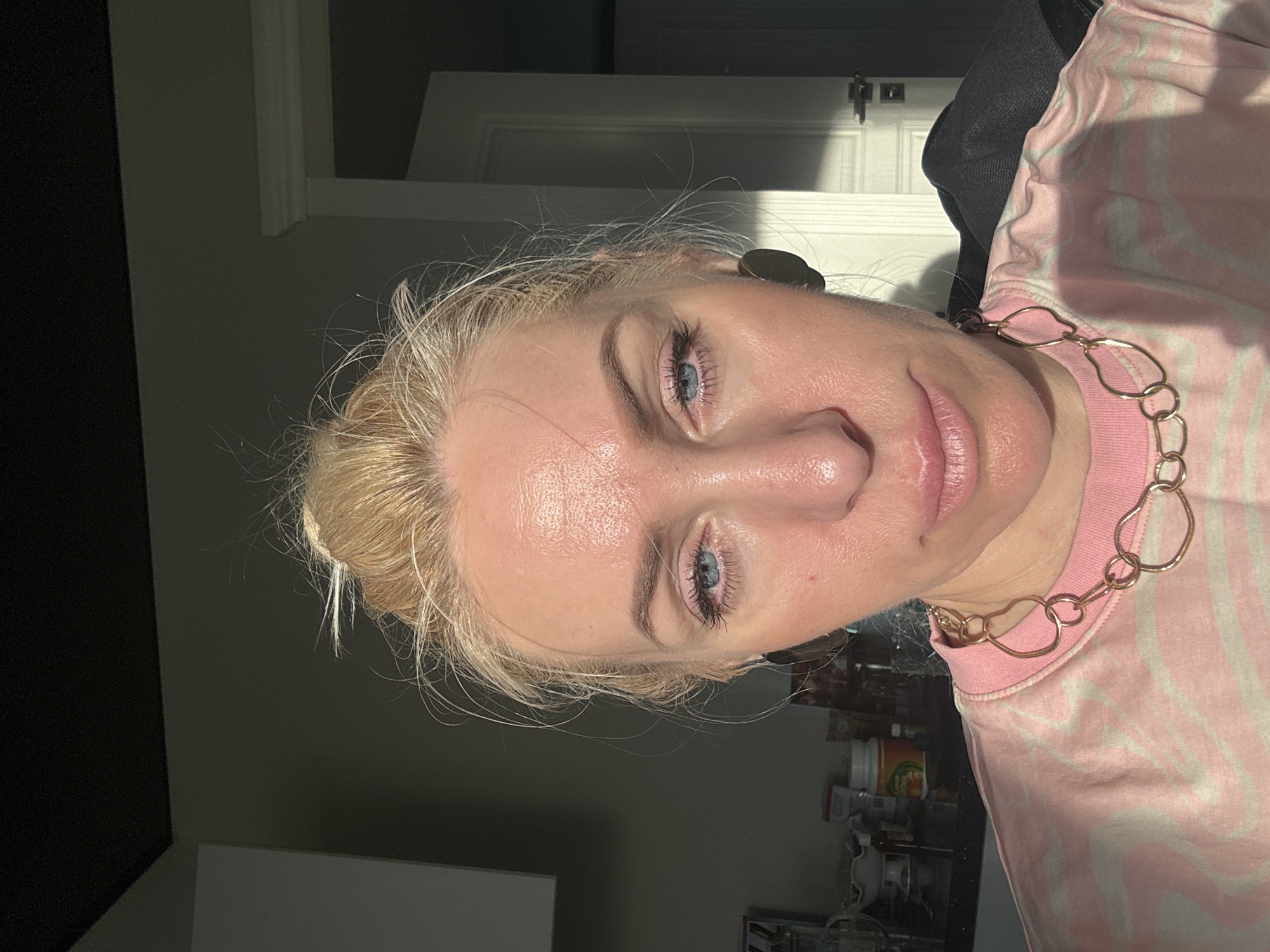 A blonde woman is wearing a pink and white patterned shirt and a gold necklace. She has blue eyes and appears to be indoors. The environment includes a door in the background and miscellaneous items visible in the lower right corner. The location is Unknown, Unknown.