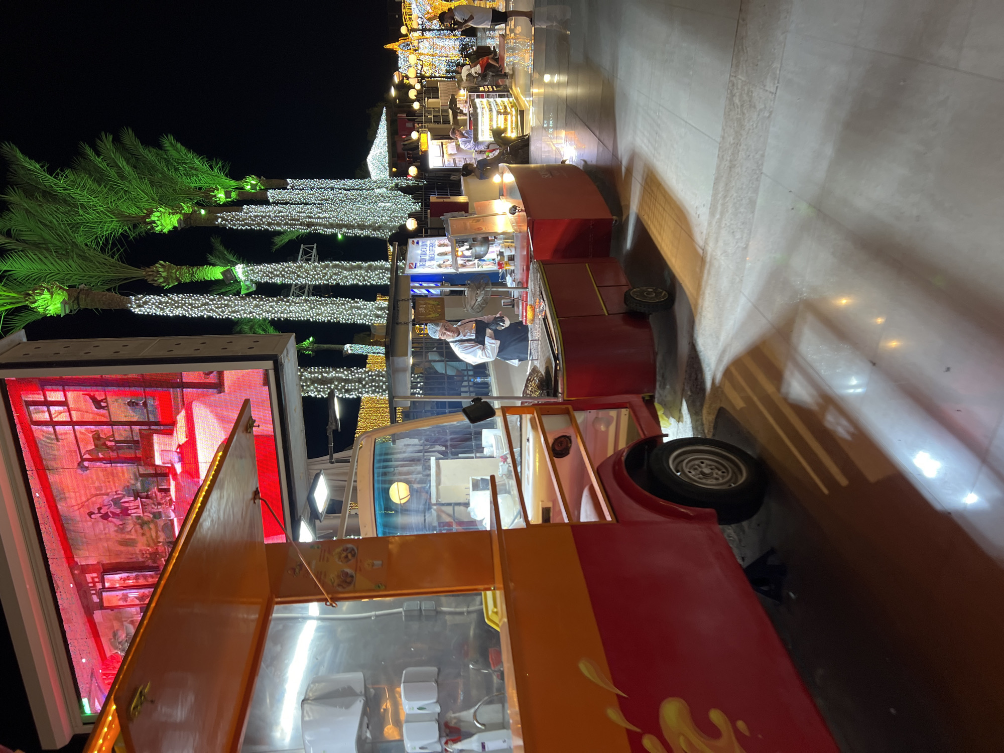 At night in Sharm El Sheikh, Egypt, a vibrant scene unfolds with a food truck as the central element. The truck is brightly lit, featuring an orange and red exterior with yellow flame-like graphics on its side. Above the food truck, a large LED screen displays an image of an interior scene with red lighting.
To the right of the food truck, a man in a white chef's coat and a black apron is actively working at a food stall. He is surrounded by various food preparation equipment, including a fan. In front of him, red food carts are set up. Further in the background, more food stalls and shops are visible, adorned with festive white and green string lights, particularly on palm trees. The ground is a tiled walkway with reflections from the lights. Several people are present, some interacting with the stalls, others walking by. The overall ambiance is that of a lively night market or promenade.