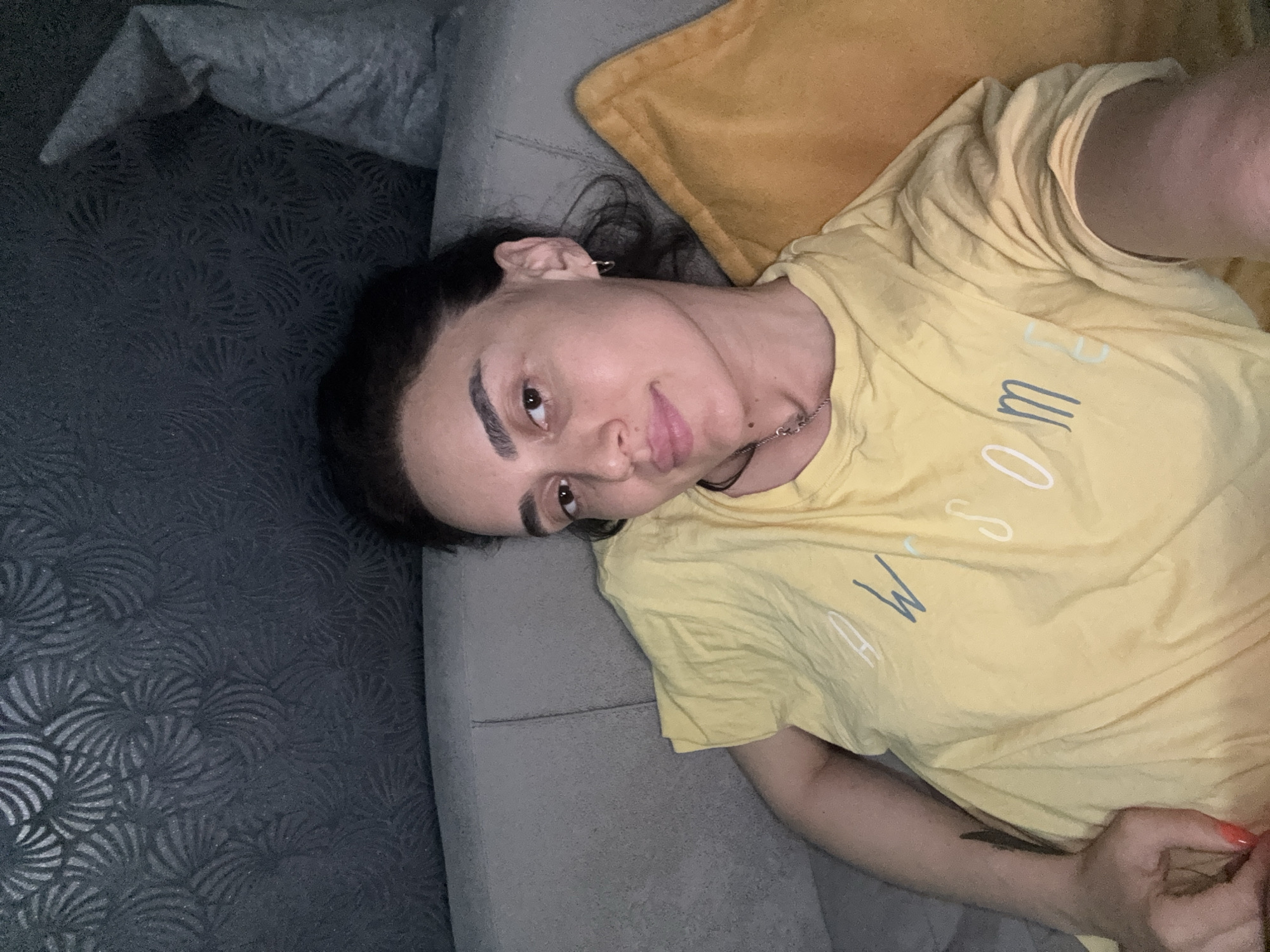 A young woman is lying on a sofa. She is wearing a yellow t-shirt with the word "AWESOME" written on it. The sofa is light gray, and behind it is a dark gray wall with a textured pattern. A yellow pillow is on the sofa next to her. The woman has dark hair and is looking at the camera. The scene appears to be indoors.
