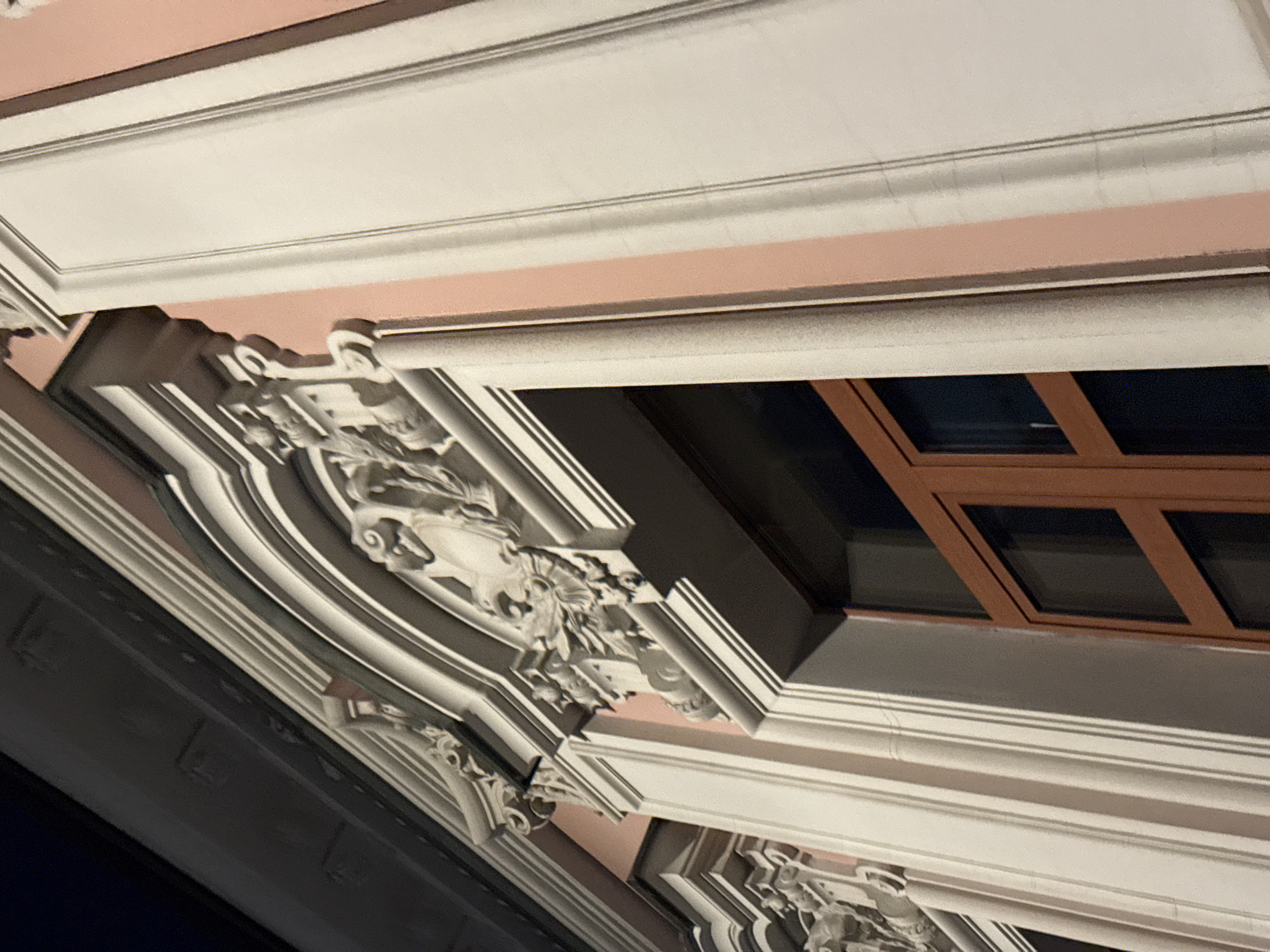 The image displays an upward-angled, close-up view of an ornate building facade in Moscow, Russia. The scene appears to be set at night or during the evening, evidenced by the dark windowpanes and the artificial lighting that highlights the architectural details.
The facade is characterized by prominent, multi-layered cornices painted in a light, almost white, color. These contrast with the soft, muted pink or peach tone of the building's wall surfaces visible behind the decorative elements. Intricate carvings, resembling grotesque masks, mythological figures, and elaborate floral or scrollwork motifs, adorn the structures beneath the cornices, showcasing detailed craftsmanship.
A window with dark panes and a distinctive brown frame is visible on the right side of the image, set within the facade. The lighting creates strong highlights and shadows, emphasizing the three-dimensional quality and texture of the architectural elements. There are no people, visible activities, or discernible text in the image.