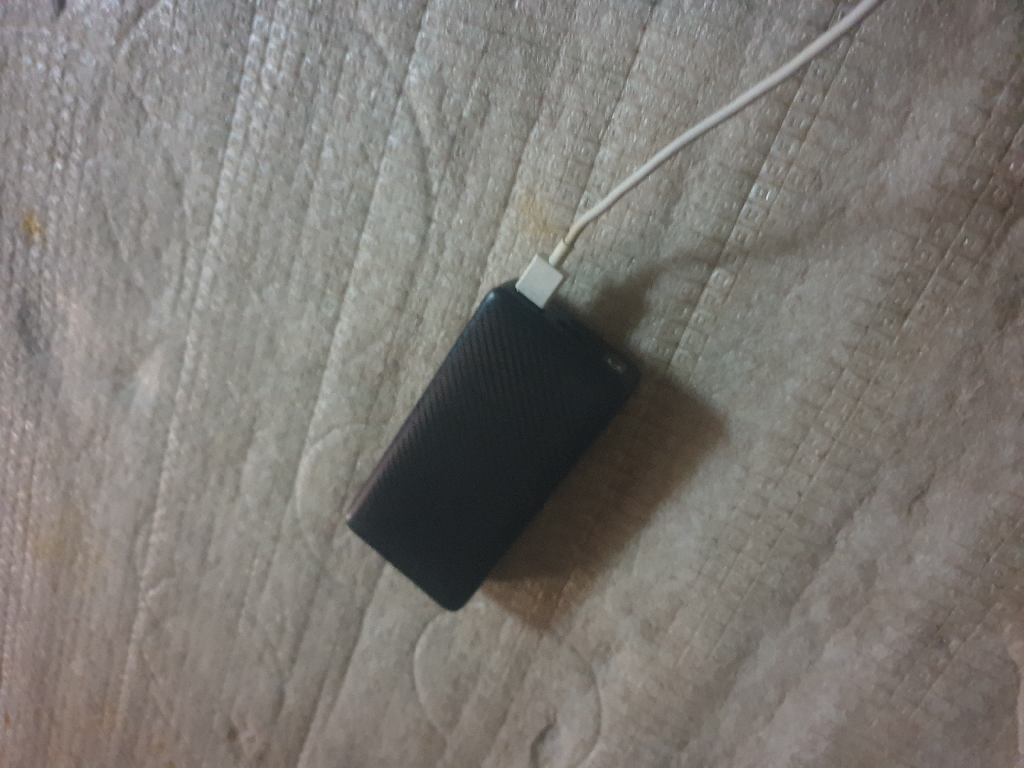 A black power bank with a white charging cable is lying on a textured, beige surface that appears to be a mattress or upholstered furniture. The power bank is positioned diagonally across the frame, with the charging cable extending from its right side and trailing off to the right. The surface has a quilted or patterned texture with visible stitching or indentations. There are no people or other objects clearly visible in the image. The lighting appears to be dim, casting a shadow from the power bank onto the surface.