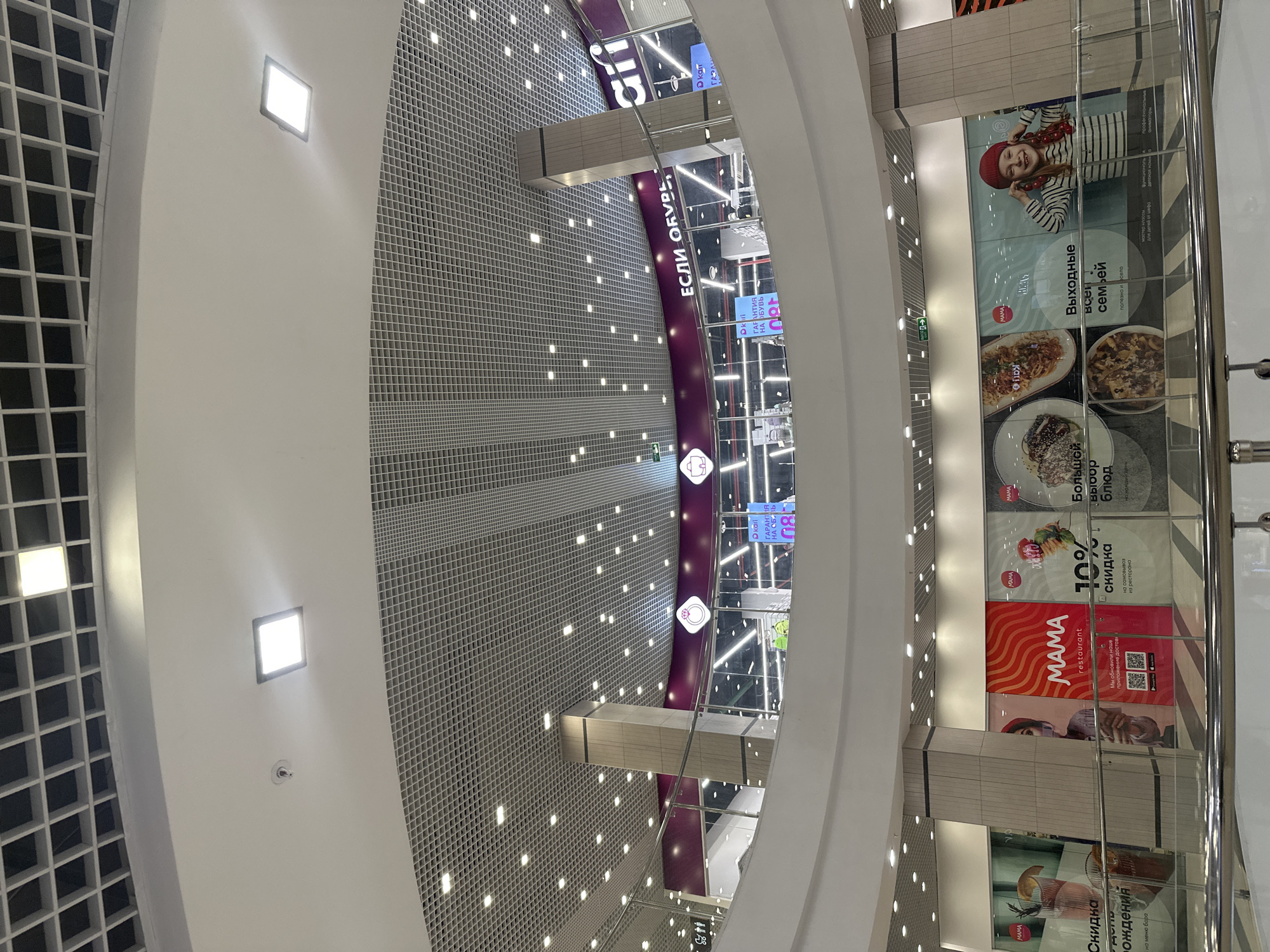 The image shows the interior of a shopping mall in Yekaterinburg, Russia. The view looks downwards from a high vantage point. The ceiling has a grid pattern with many small square lights embedded in it. A curved, dark purple band runs through the center of the frame, marked with shop names and logos, including "Kari" and "Если Обувь". A glass barrier lines the right side of the image, displaying advertisements with images of food, people, and the word "Mama" prominently featured. The overall impression is of a modern shopping center interior.