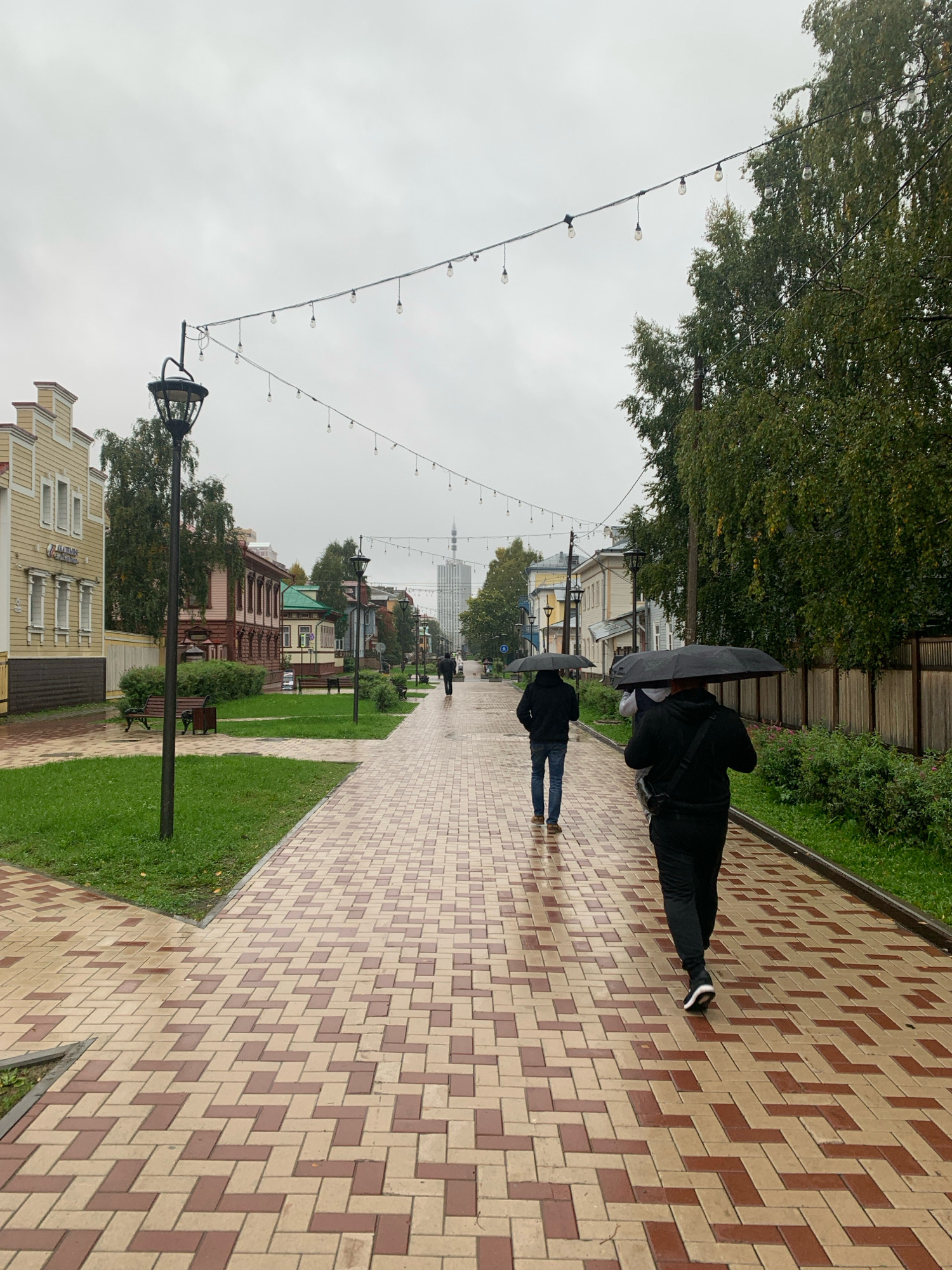 A street scene in Arkhangelsk, Russia, on a cloudy, wet day. The foreground features a brick-paved walkway with a pattern of red and beige bricks. To the left, a strip of green grass with benches and a lamppost is visible, adjacent to a light-yellow building. Further down the street, traditional Russian wooden houses with colorful roofs line the path, interspersed with trees. In the background, a tall, modern skyscraper stands against the overcast sky. Several people are walking on the path, all holding black umbrellas, indicating rain. Two individuals are prominently in the foreground, walking away from the camera. The overall atmosphere is subdued due to the weather.