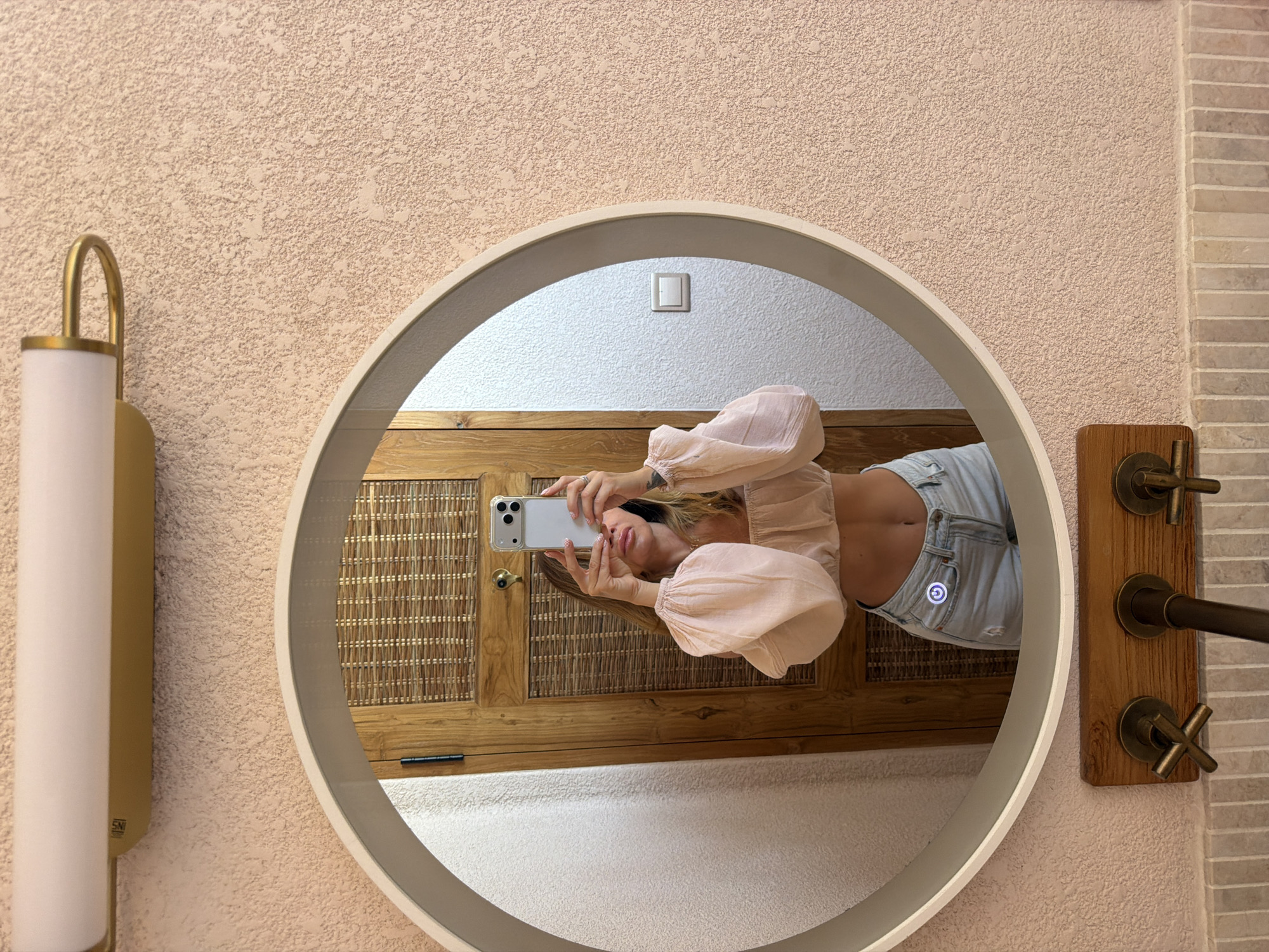 A woman stands in front of a round mirror, taking a selfie. She is wearing a light pink off-the-shoulder crop top with voluminous sleeves and light blue denim shorts. The mirror reflects her and a wooden door with a woven inset behind her. The wall in the background is a textured pink. A light fixture with a gold frame is mounted above the mirror. Below the mirror are two vintage-style brass faucets mounted on a wooden panel. The location is identified as Mengwi, Indonesia.